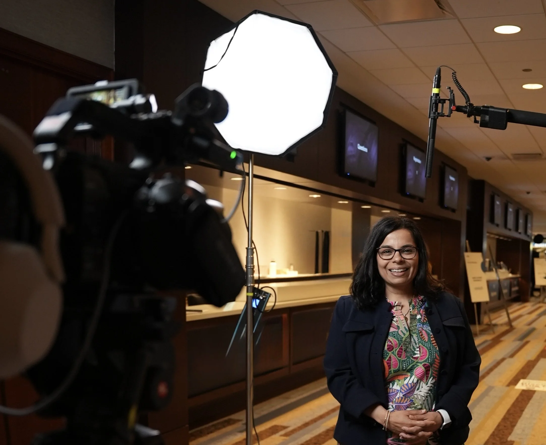 A woman with glasses and dark hair, smiling, standing in front of a camera and bright light, in a conference or event setting, with monitors on the wall behind her.