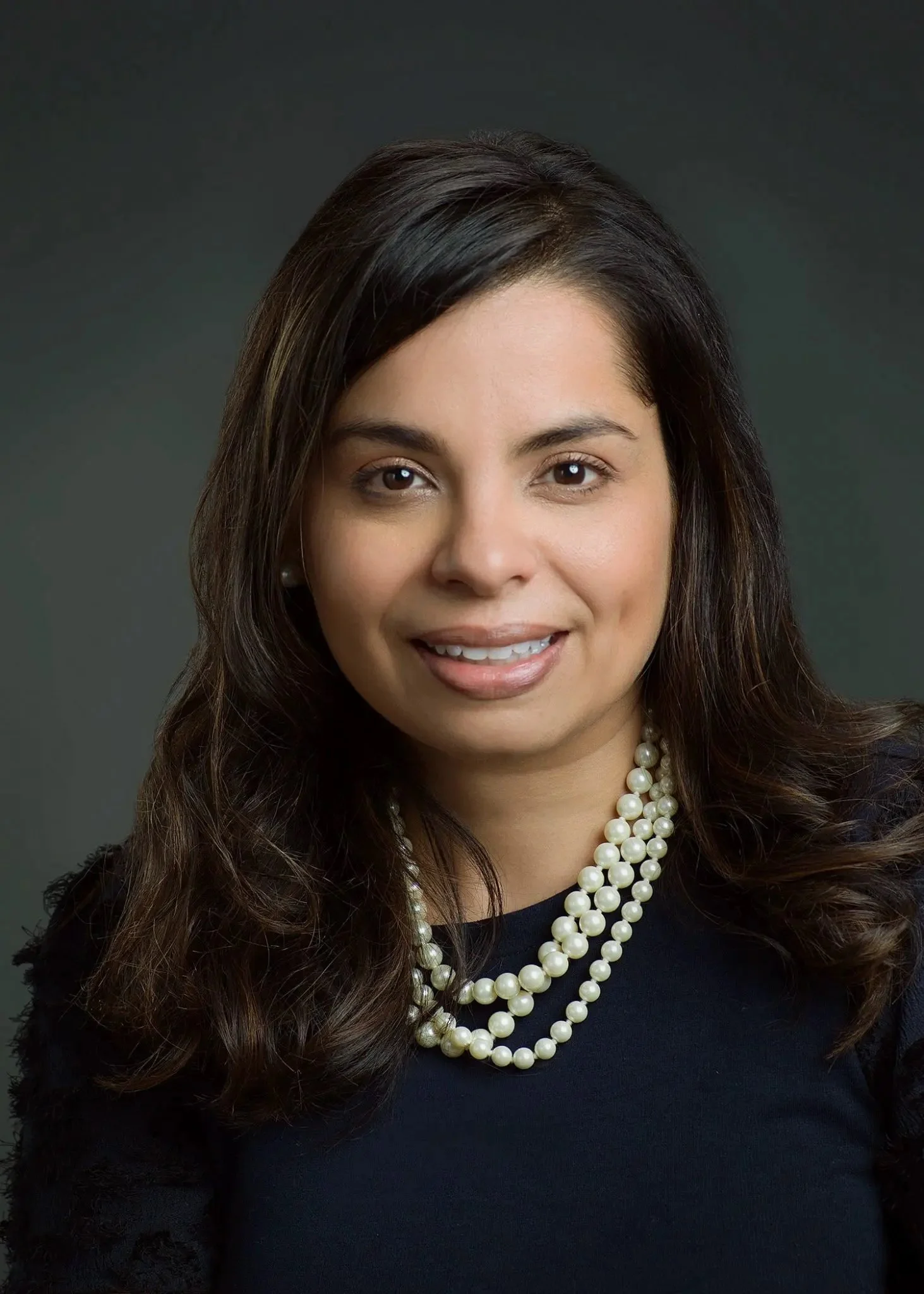 A woman with dark hair and a pearl necklace smiling in a portrait photo.