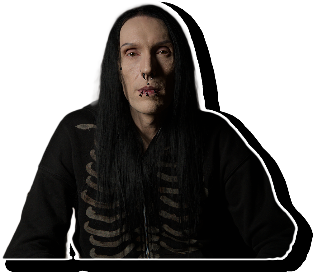 A person with long black hair, pale skin, and facial piercings, wearing a black hoodie with a skeleton design on the front, sitting against a black background.