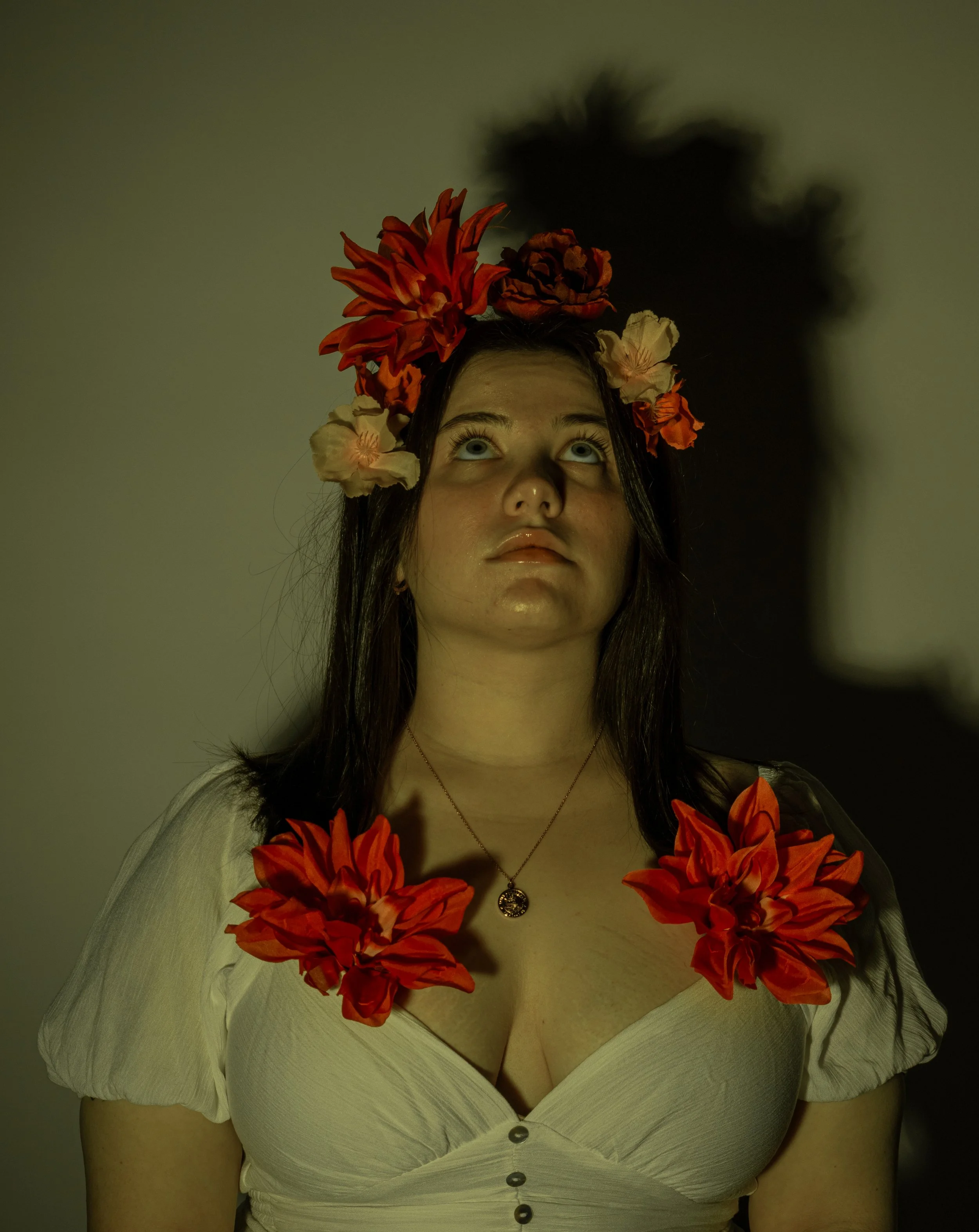 A woman with a floral headpiece and flowers on her shoulders, looking upward with a shadow cast on the wall behind her.
