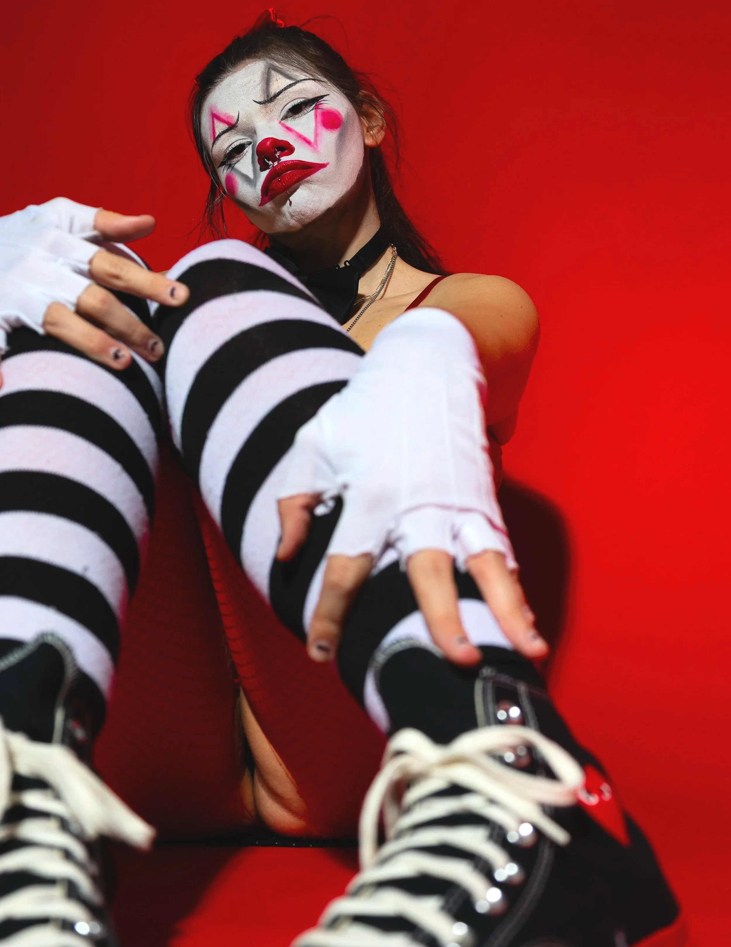 Person dressed as a clown with white face paint, red nose, and colorful makeup, wearing striped stockings, black shoes, and fingerless gloves, sitting on a red background.