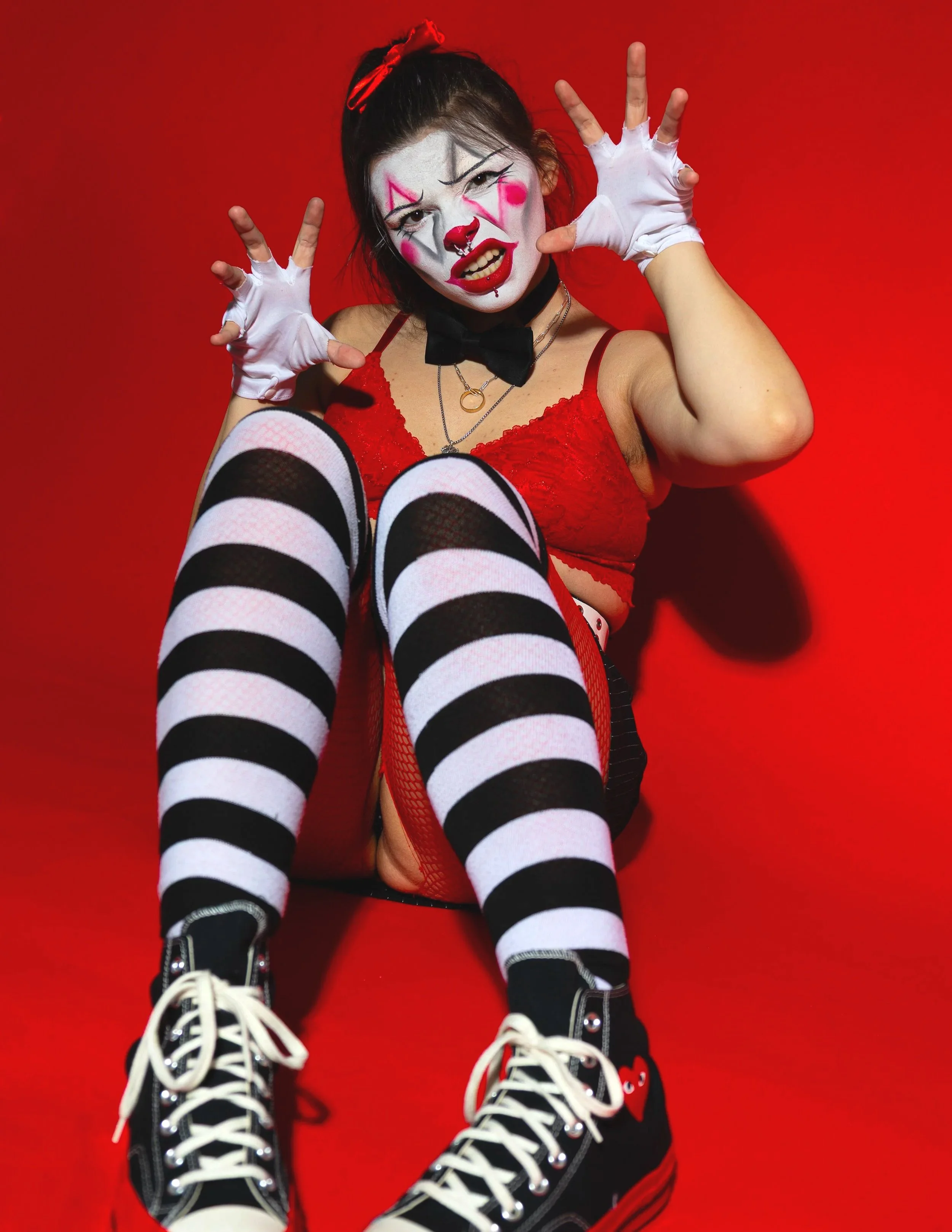 Person dressed as a clown in red lingerie, black and white striped thigh-high socks, black Converse sneakers, with clown makeup on face, posing against red background.