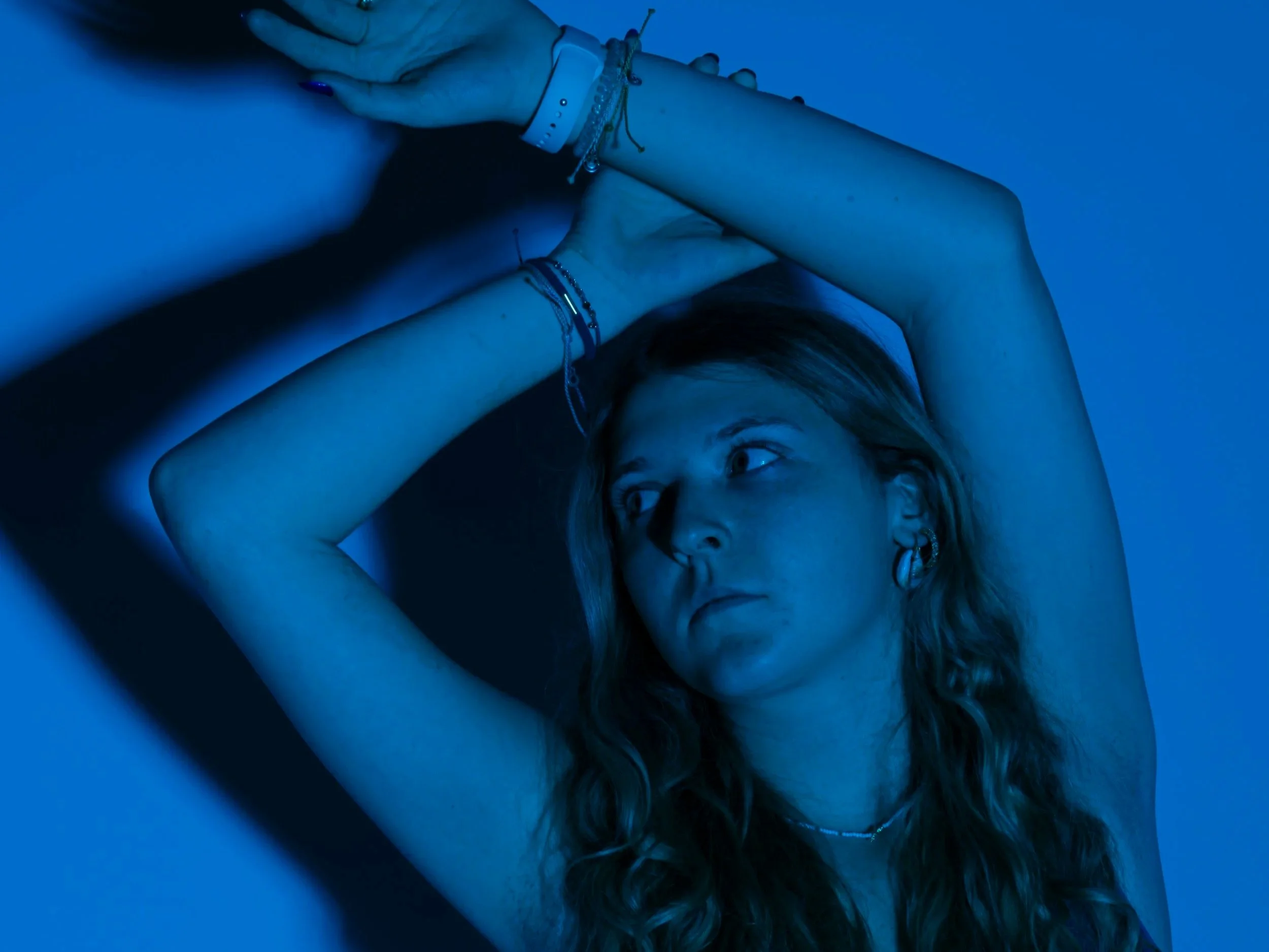 A young woman with long curly hair, earrings, and bracelets, gazing to the side with her arms raised above her head, illuminated by blue light.