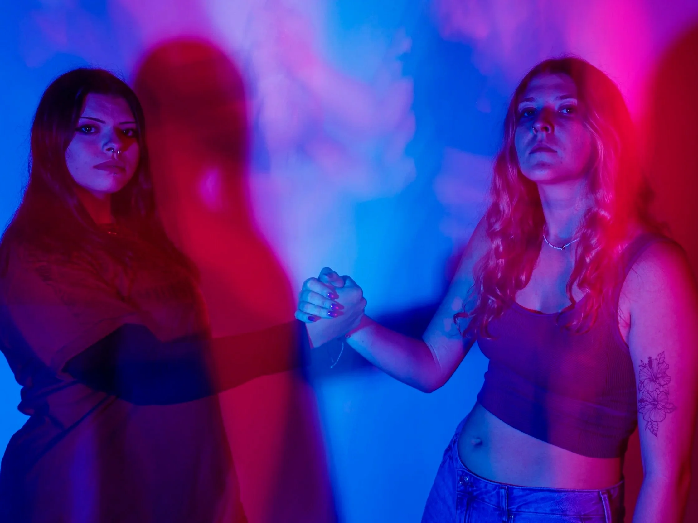 Two women with long hair holding hands, illuminated by pink and blue neon lights.