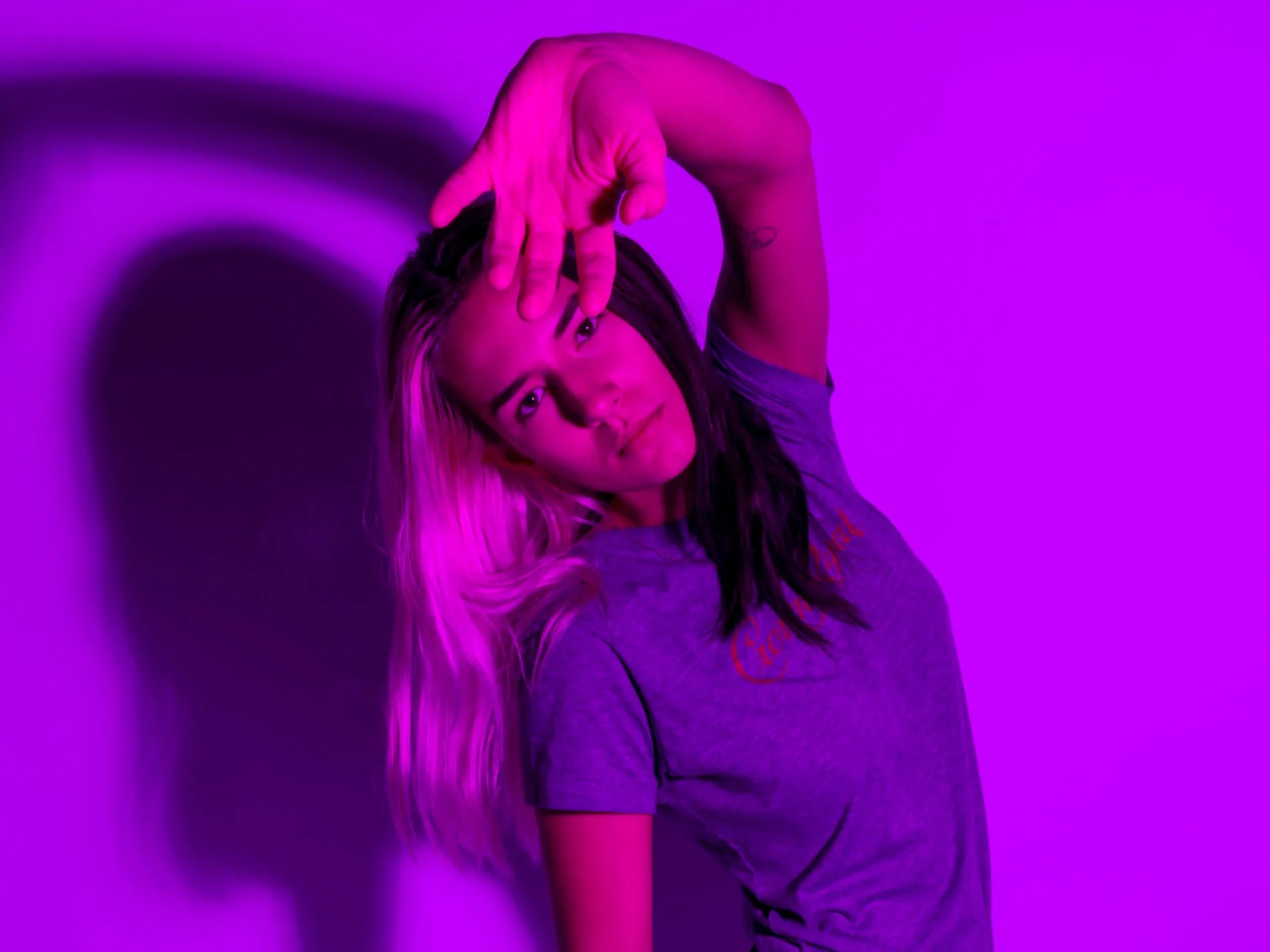 A woman with long dark hair, wearing a gray t-shirt, poses against a purple background with dramatic lighting and shadow.
