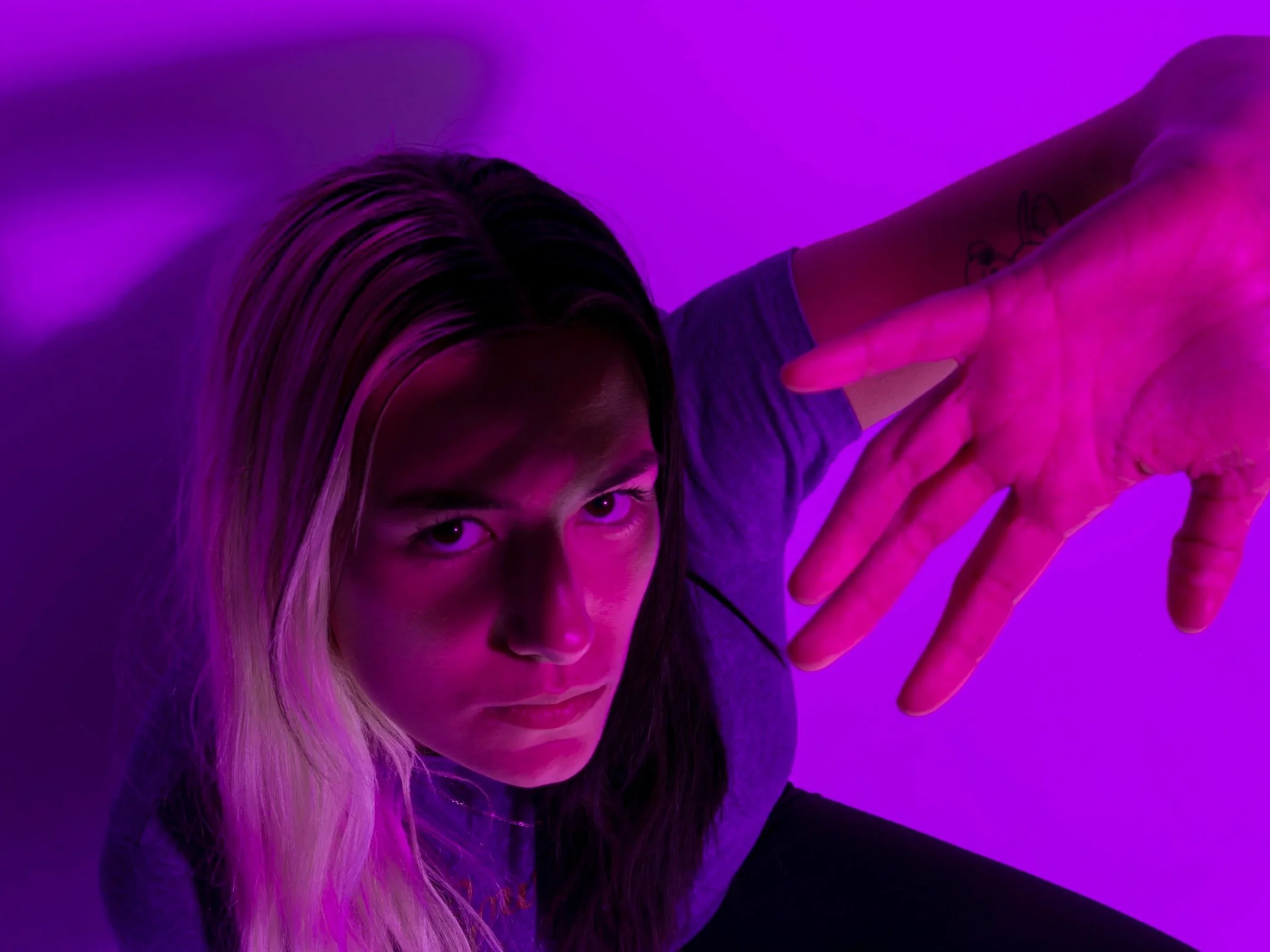 A woman with long blonde hair looking up at the camera, illuminated by purple-pink lighting, with her hand extended toward the camera in a dramatic pose.