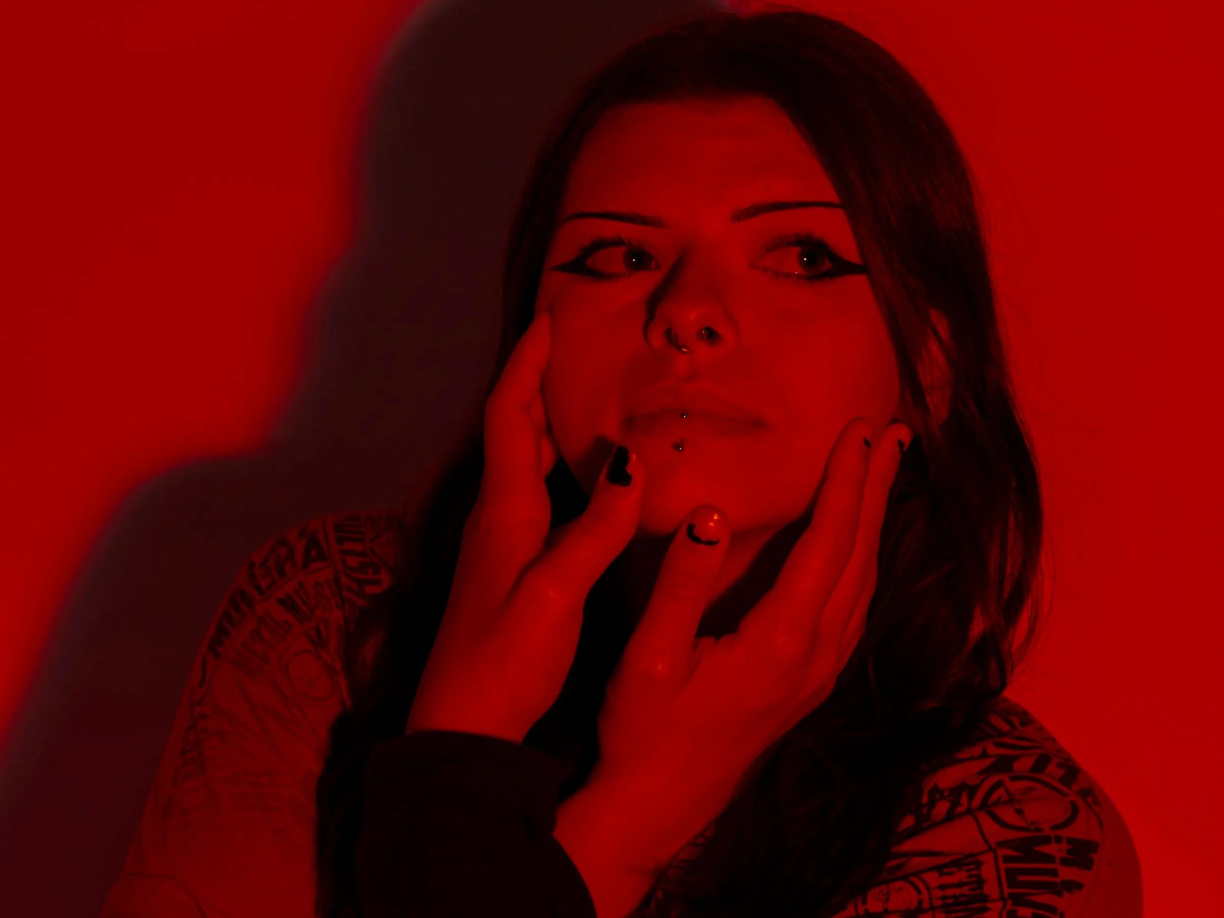 Close-up of a woman with dark hair, multiple facial piercings, and tattoos, touching her face with both hands, in a dim red lighting environment.