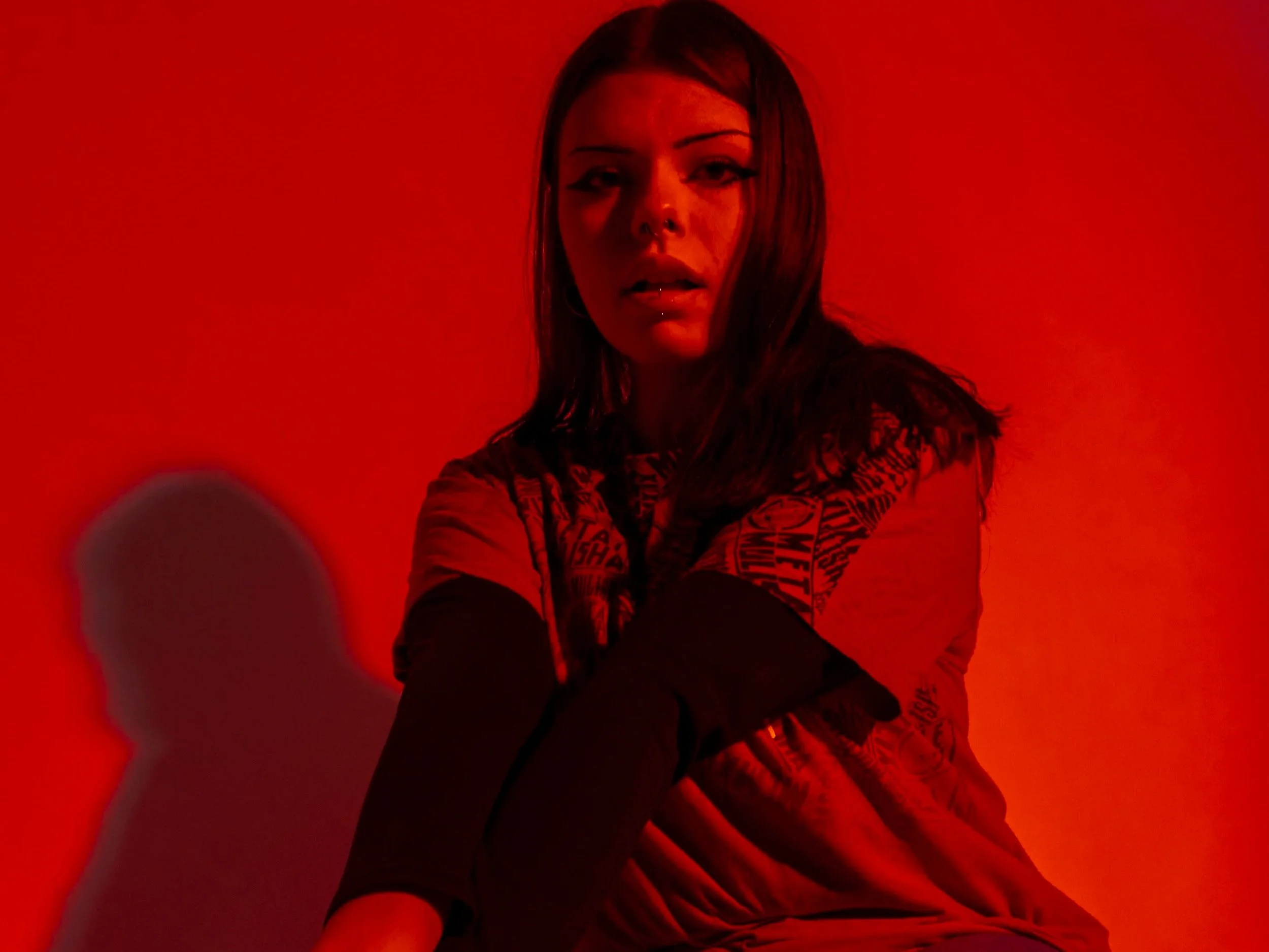 A woman with dark hair and makeup sitting against a red background, casting a shadow on the wall.