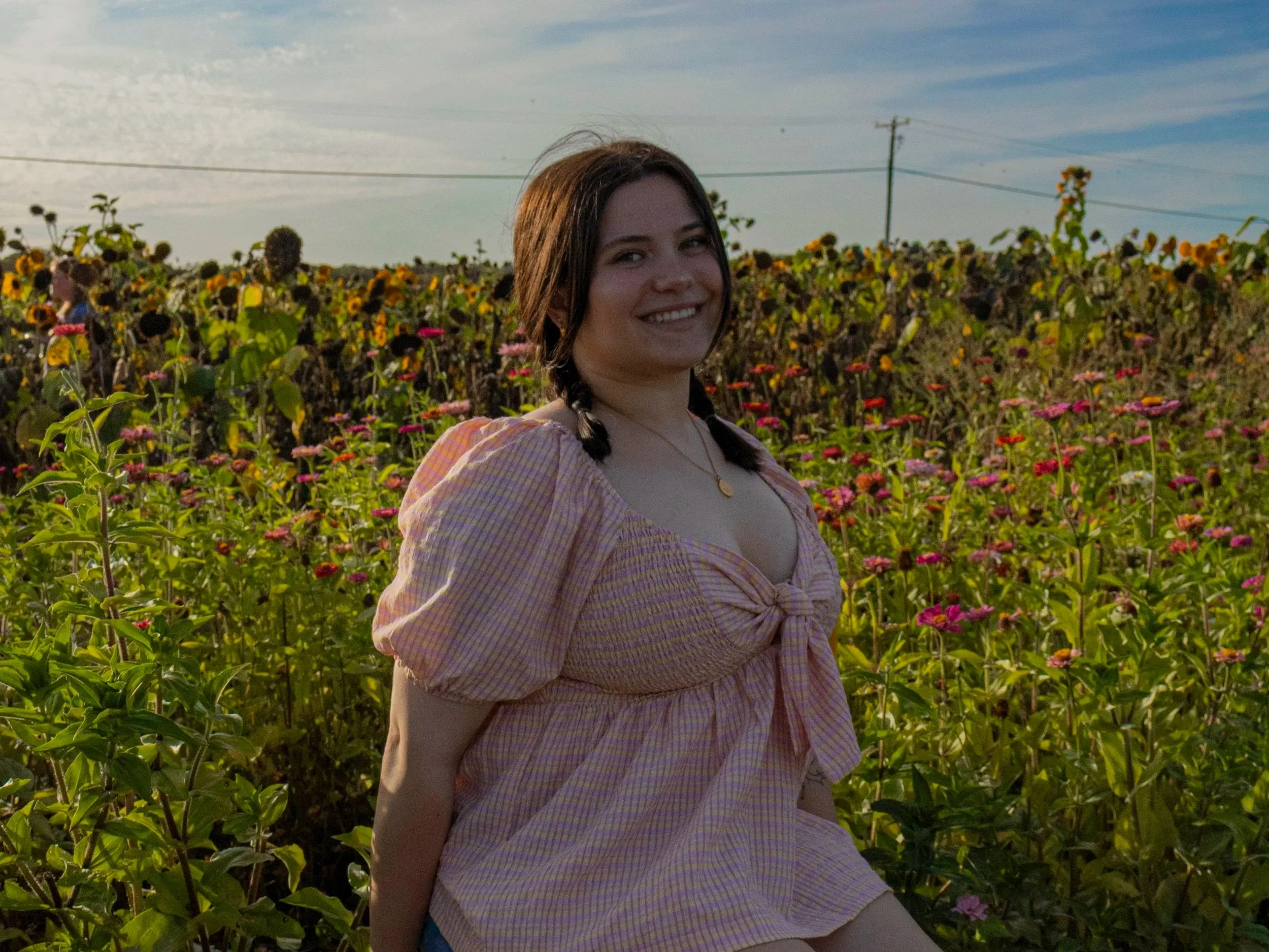 A young woman with dark braided hair and a necklace, smiling while sitting in a field of blooming flowers at sunset.