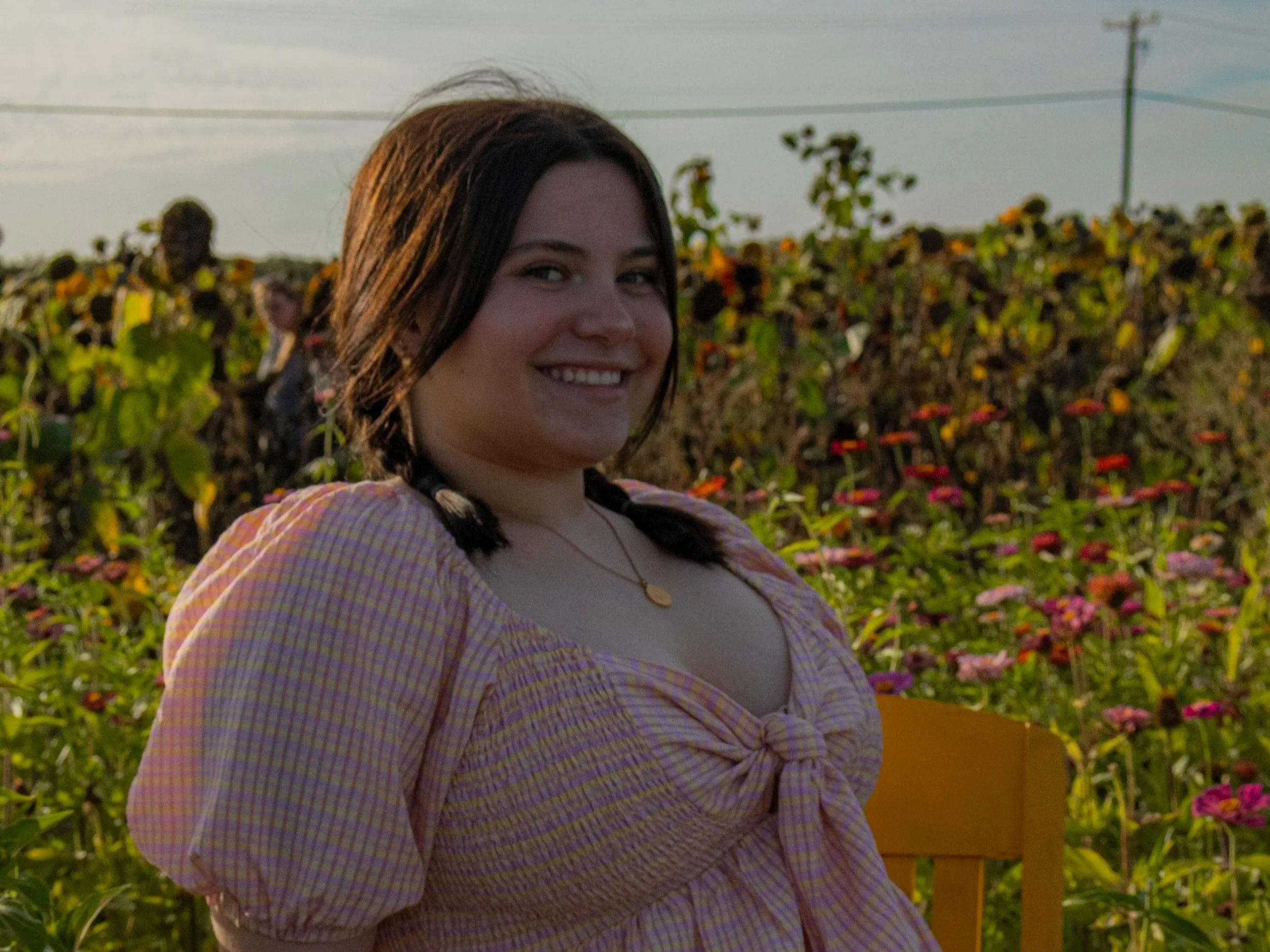 A smiling young woman with dark hair styled in two braids, wearing a pink checkered dress with puffed sleeves, sitting outdoors in a garden with colorful flowers in the background during sunset.