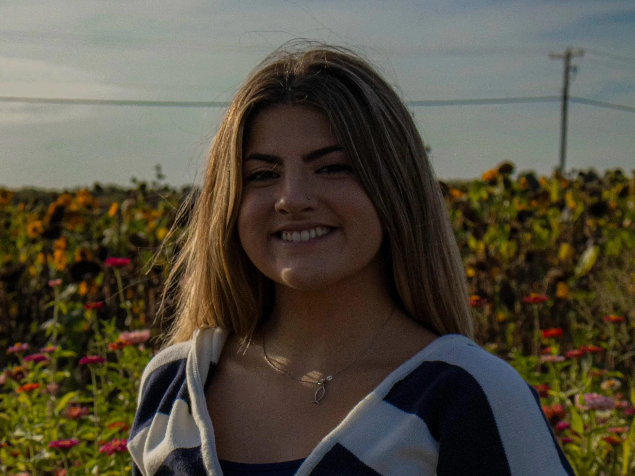 A young woman with long brown hair smiling outdoors in a field of colorful flowers during sunset.