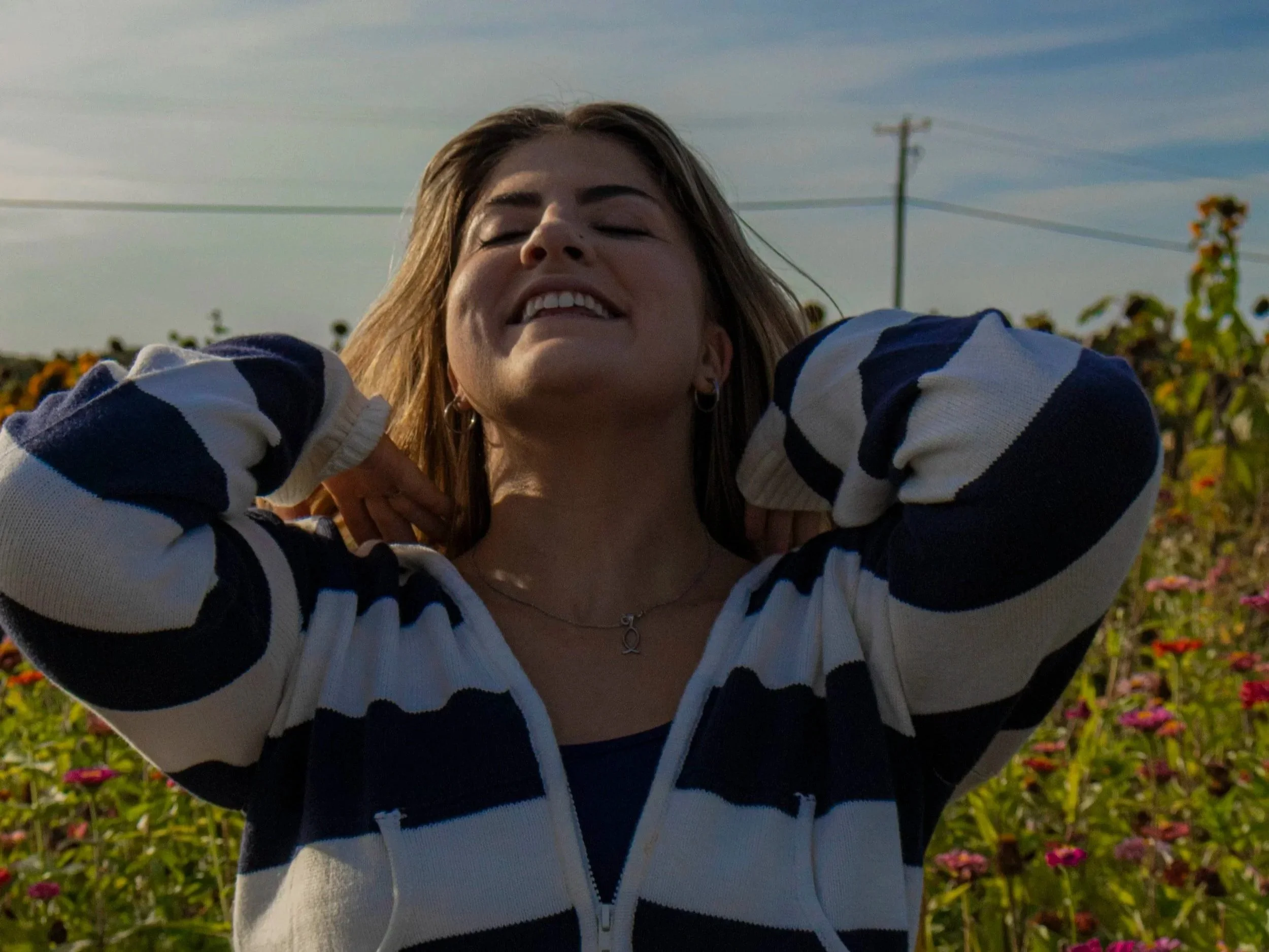 A young woman smiling and enjoying a sunny day outdoors, standing in a field of flowers with her hands behind her neck, wearing a striped navy and white jacket and a necklace.