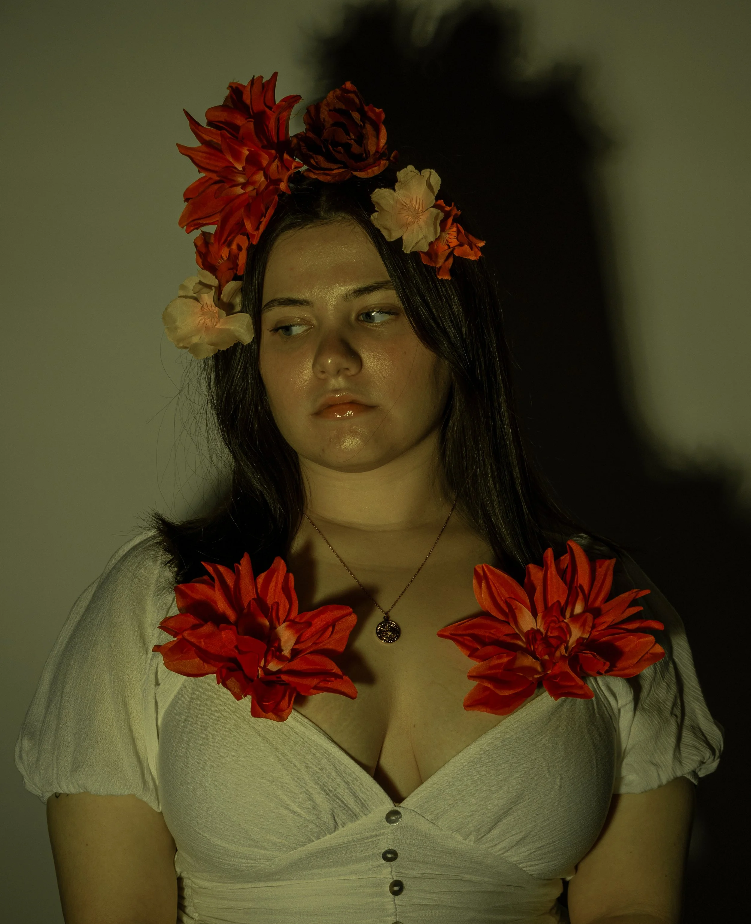 A woman with long dark hair wearing a white blouse with buttons, decorated with red and cream flowers on her head and shoulders, and a necklace with a circular pendant.