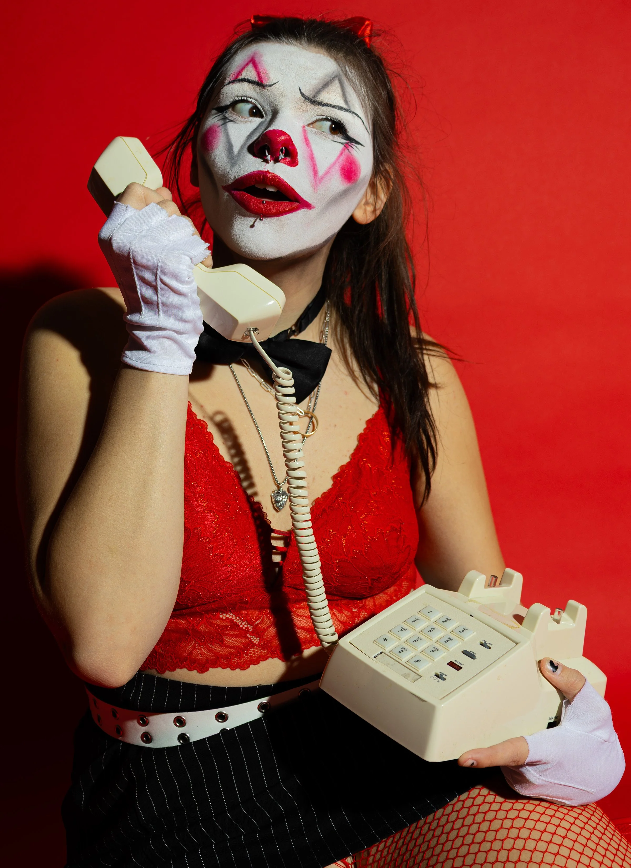 A woman with clown makeup, including a red nose and painted face, holding a vintage beige rotary phone to her ear against a red background.