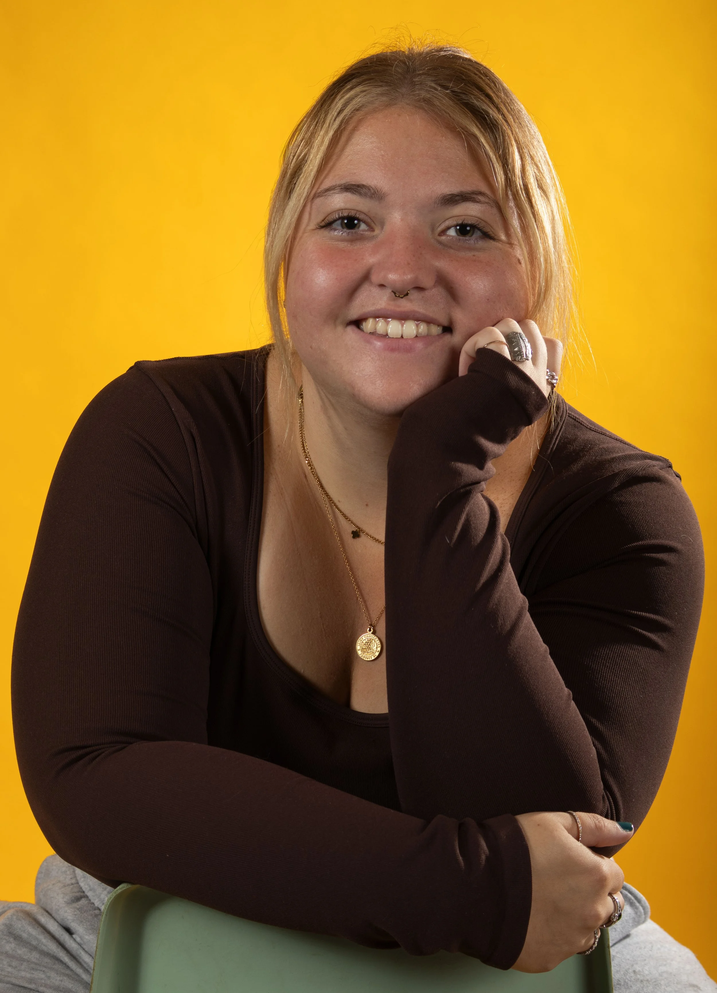 A young woman with blonde hair and a septum piercing smiling at the camera, sitting with her chin on her hand, against a bright yellow background.