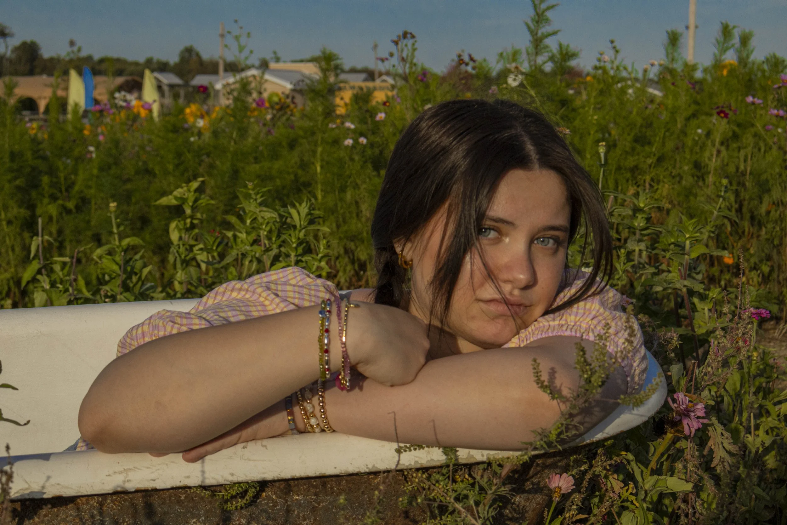 A young woman with dark hair and blue eyes lying on a white surface outdoors, surrounded by green plants and colorful flowers, wearing a pink checkered shirt and multiple bracelets.