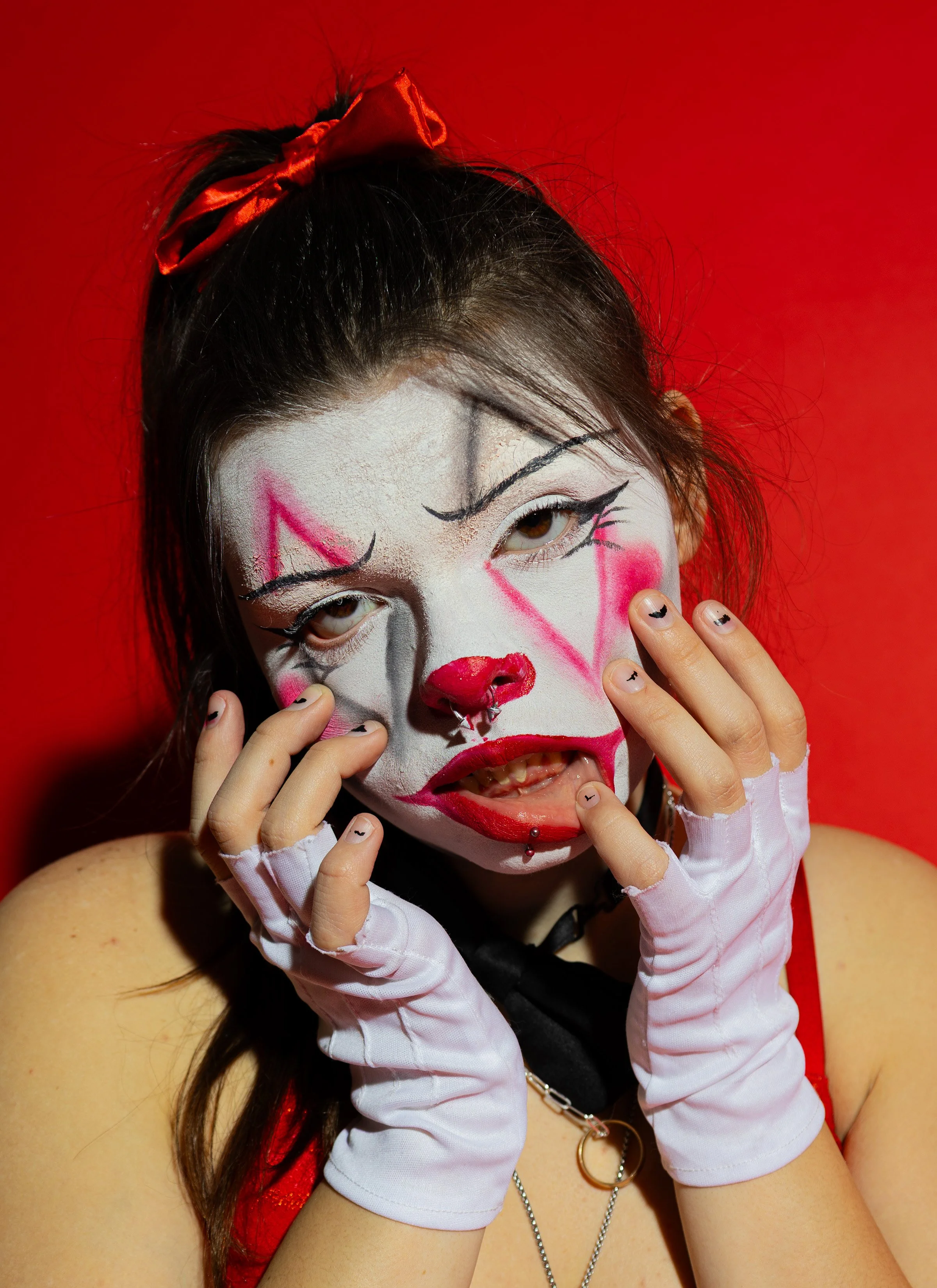 A person with clown makeup faces a bright red background, wearing white gloves, a red bow in their hair, and has facial piercings.