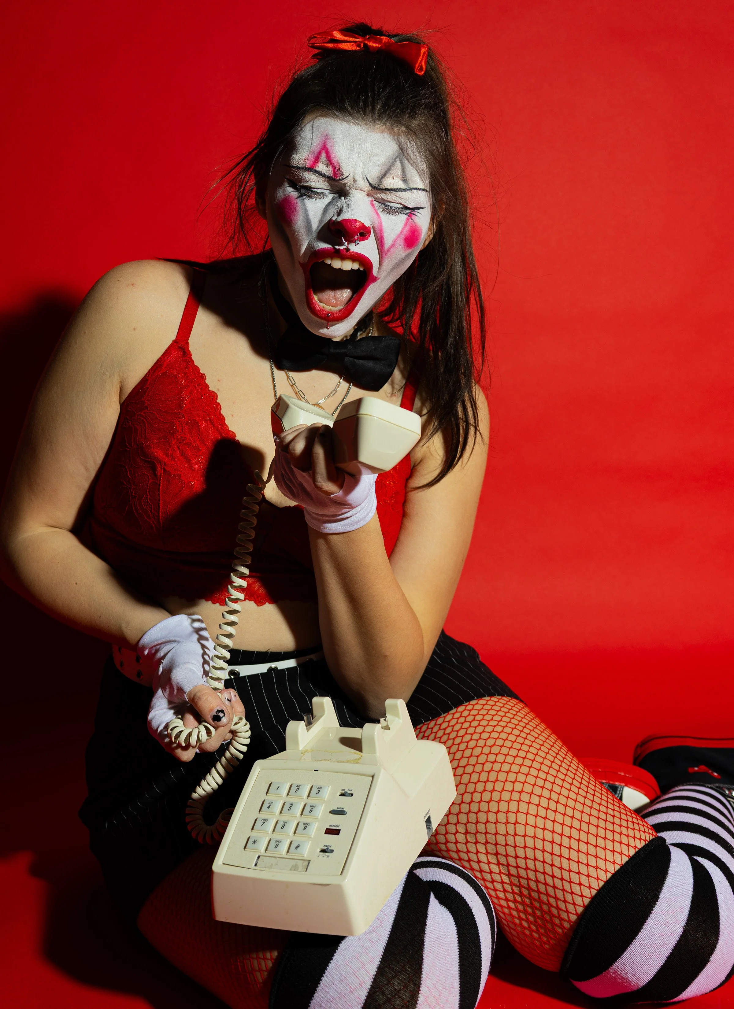 A woman dressed as a clown with face paint, wearing a red lace top, black striped shorts, red fishnet stockings, and striped socks, sitting on the floor with a vintage beige rotary dial telephone, against a red background, with her mouth open as if s