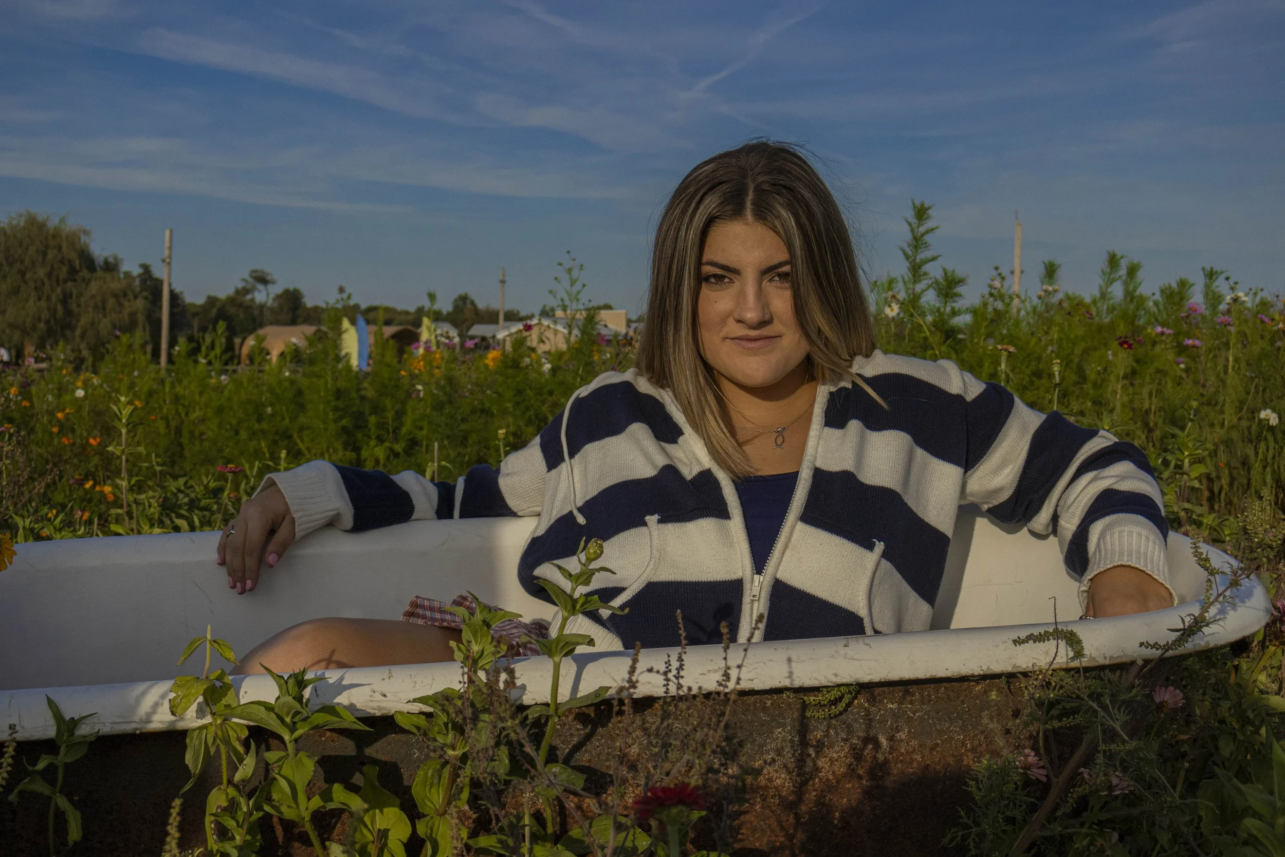 A young woman sitting in an old bathtub outdoors in a greenfield with flowers, wearing a striped navy and white hoodie, with a sunny blue sky overhead.