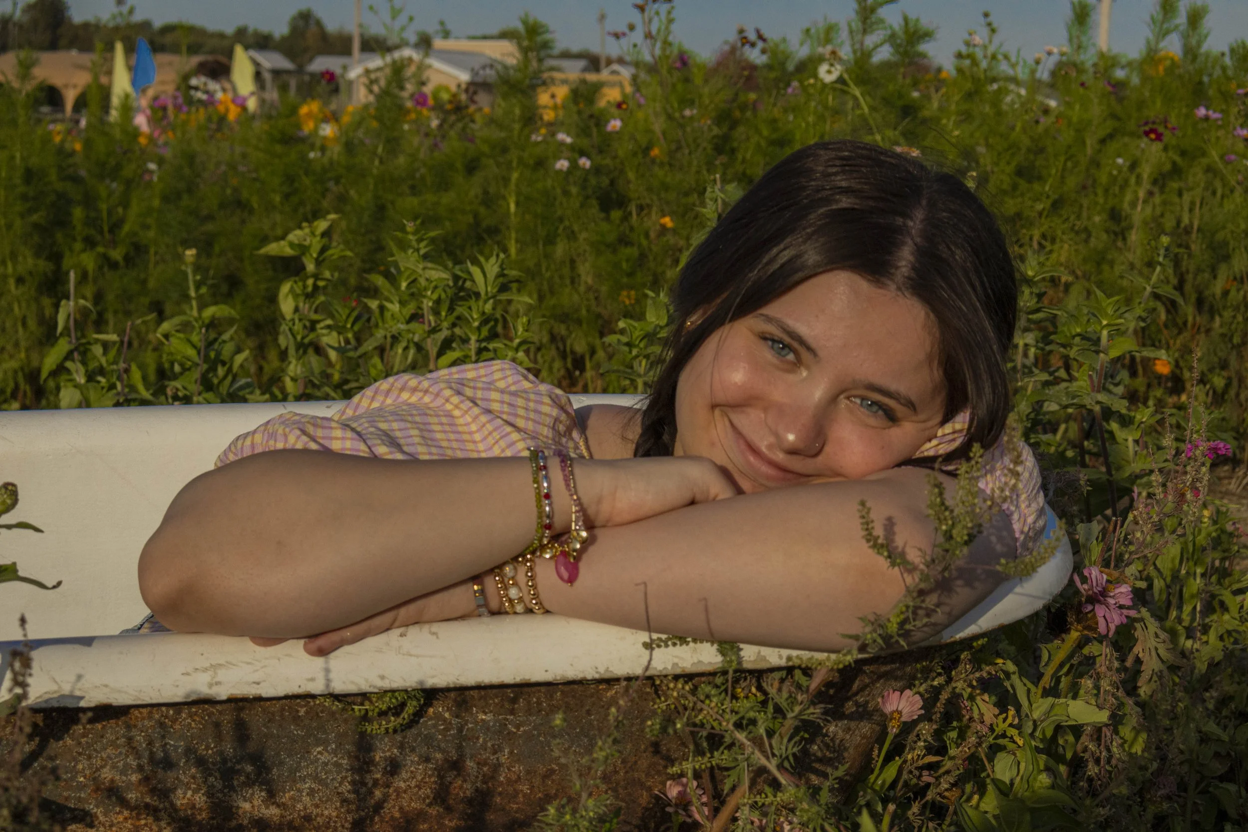 A young woman with dark hair and blue eyes lying on her side in an old bathtub outdoors, resting her head on her arms, surrounded by a field of colorful wildflowers under a clear blue sky.