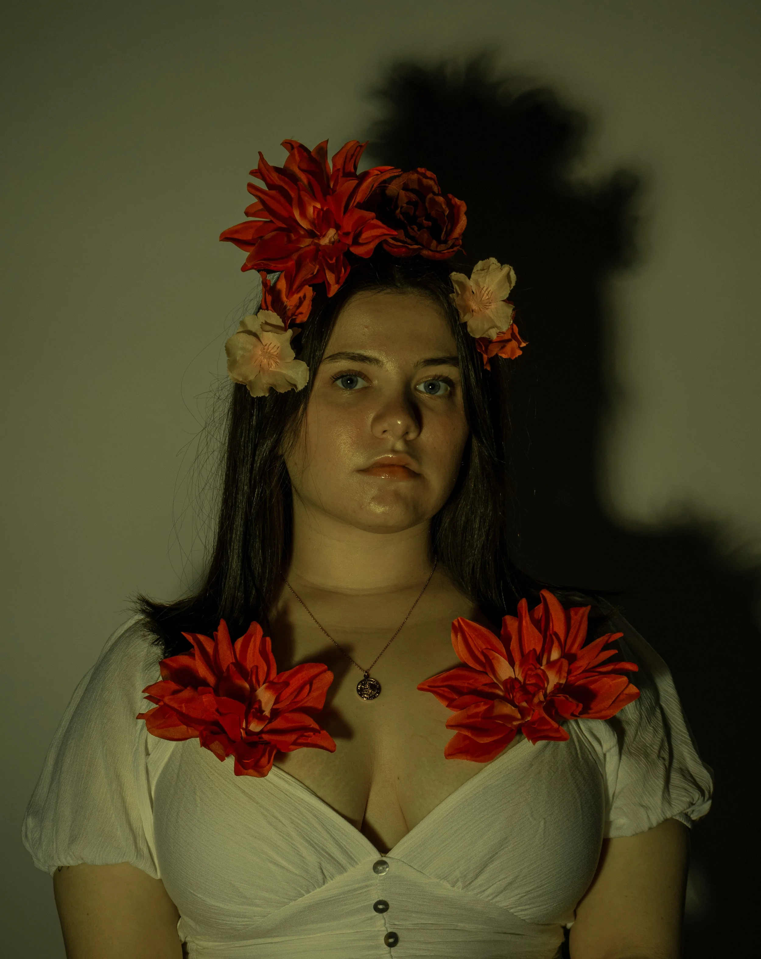 A young woman with long dark hair, wearing a white top and a necklace, is adorned with large red and pink flowers on her head and shoulders. Her shadow is cast on the wall behind her.