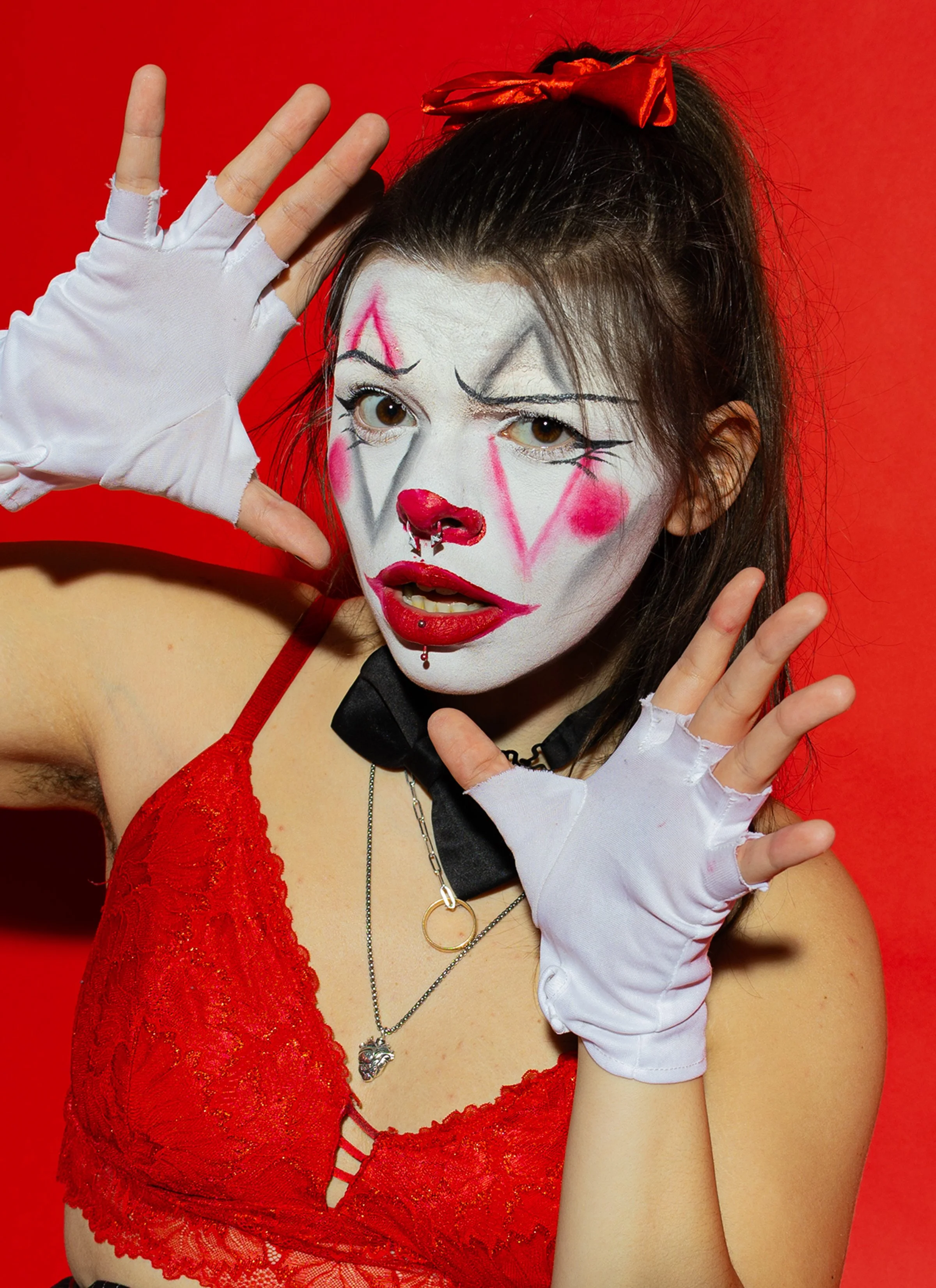 Person in clown makeup with white face paint, red and black accents, red lips, and a red nose, wearing a black bow tie and red lace top, posing against a red background with hands raised, wearing white gloves.