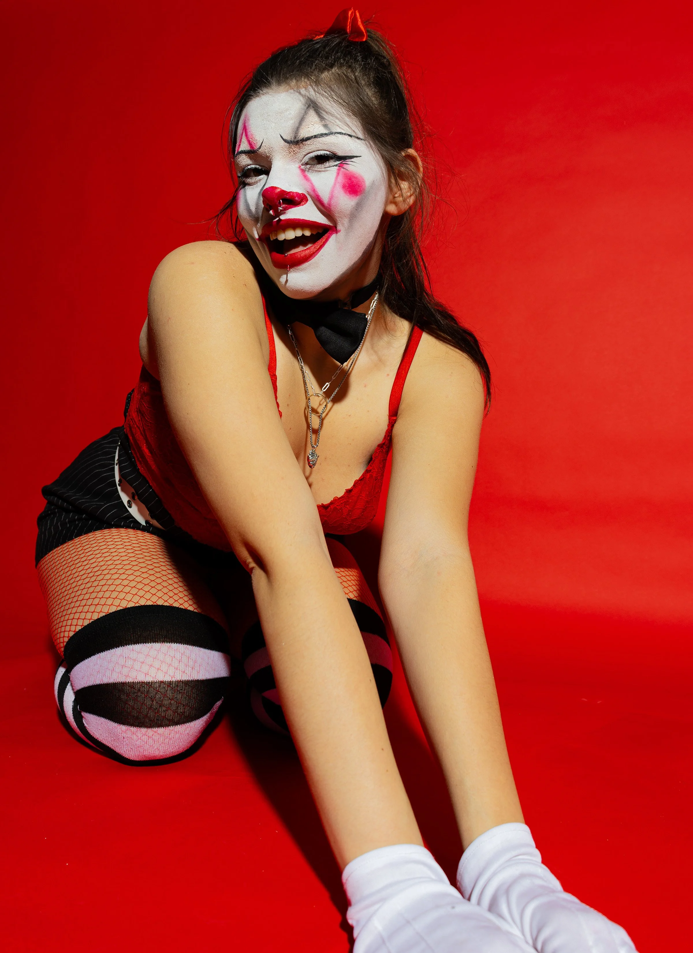 A woman dressed as a clown with white face paint, red nose, and clown makeup, smiling against a red background. She is wearing a red top, fishnet stockings, and white gloves.
