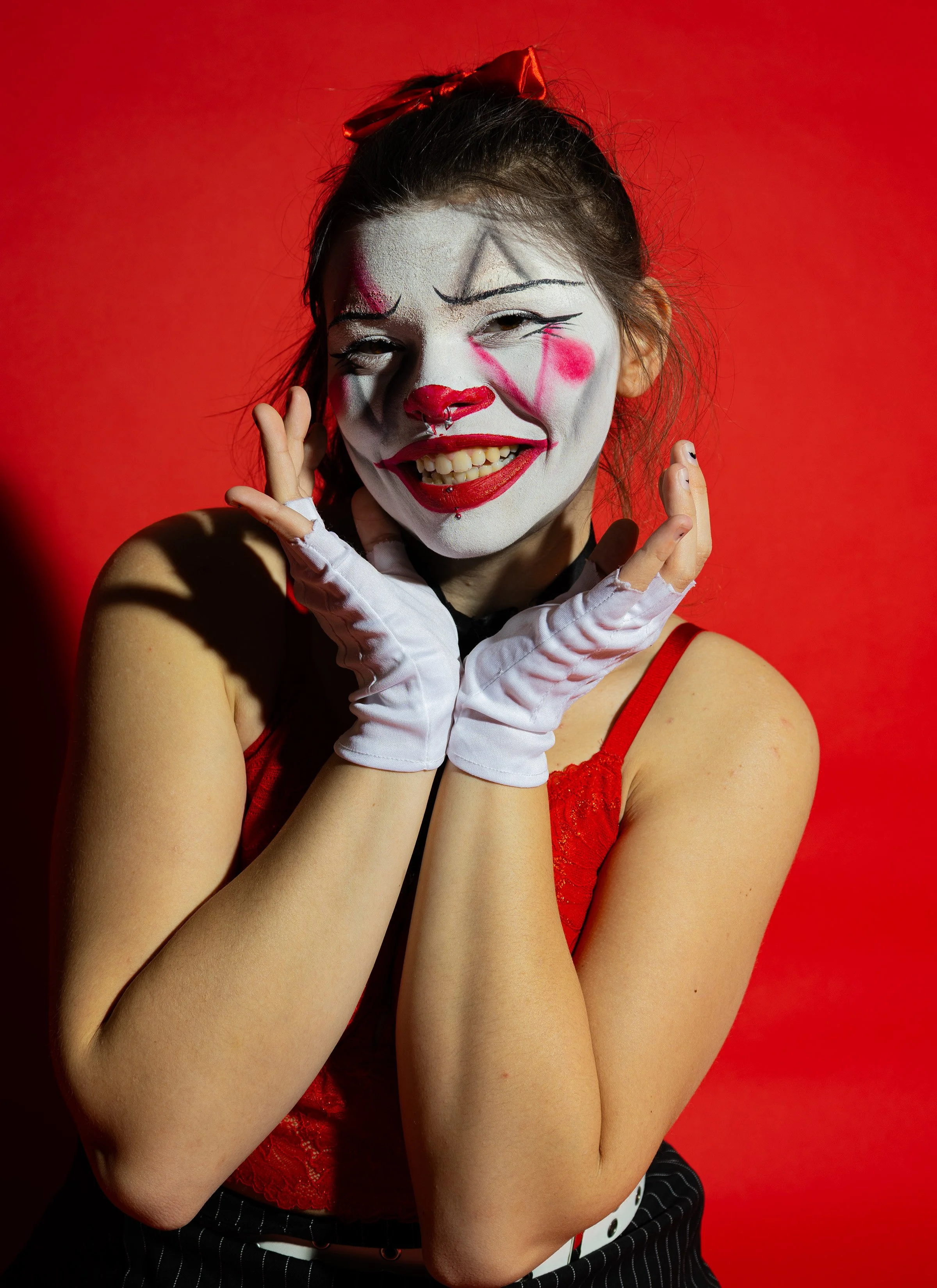 Woman dressed as a clown with white face makeup, red nose, and black and pink accents, wearing a red dress with red bow and white gloves, posing with hands near her face against a red background.