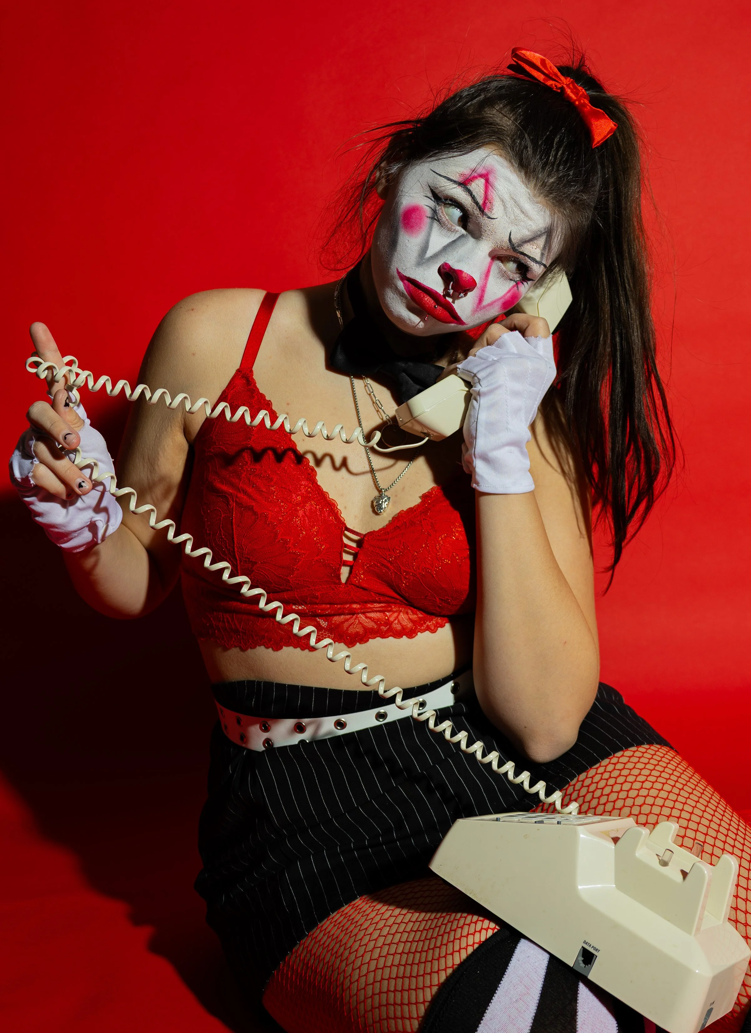 Person dressed as a clown with white face paint, red smile, and clown makeup, holding a vintage beige telephone receiver to her ear and sitting against a red background. She wears a red lace top, striped black and white shorts, fishnet stockings, whi
