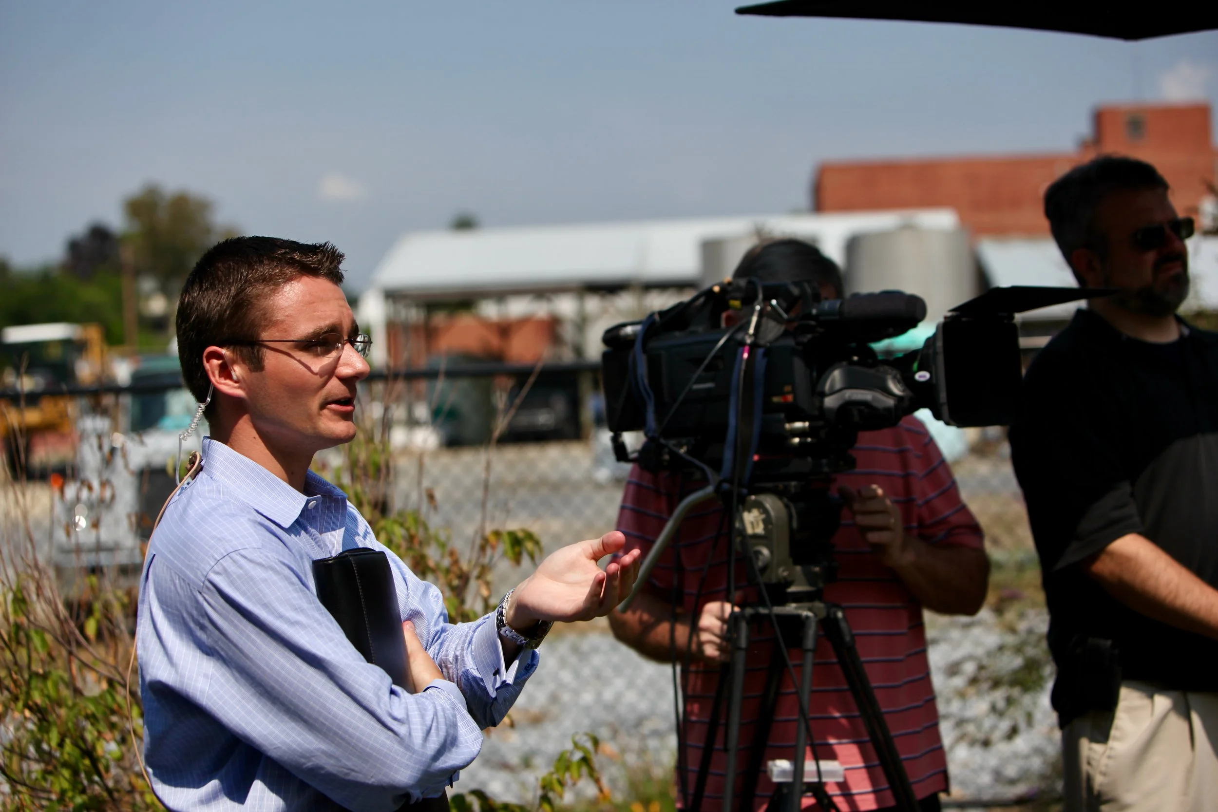 A man in a blue shirt speaking while being filmed outside with a video camera and crew.