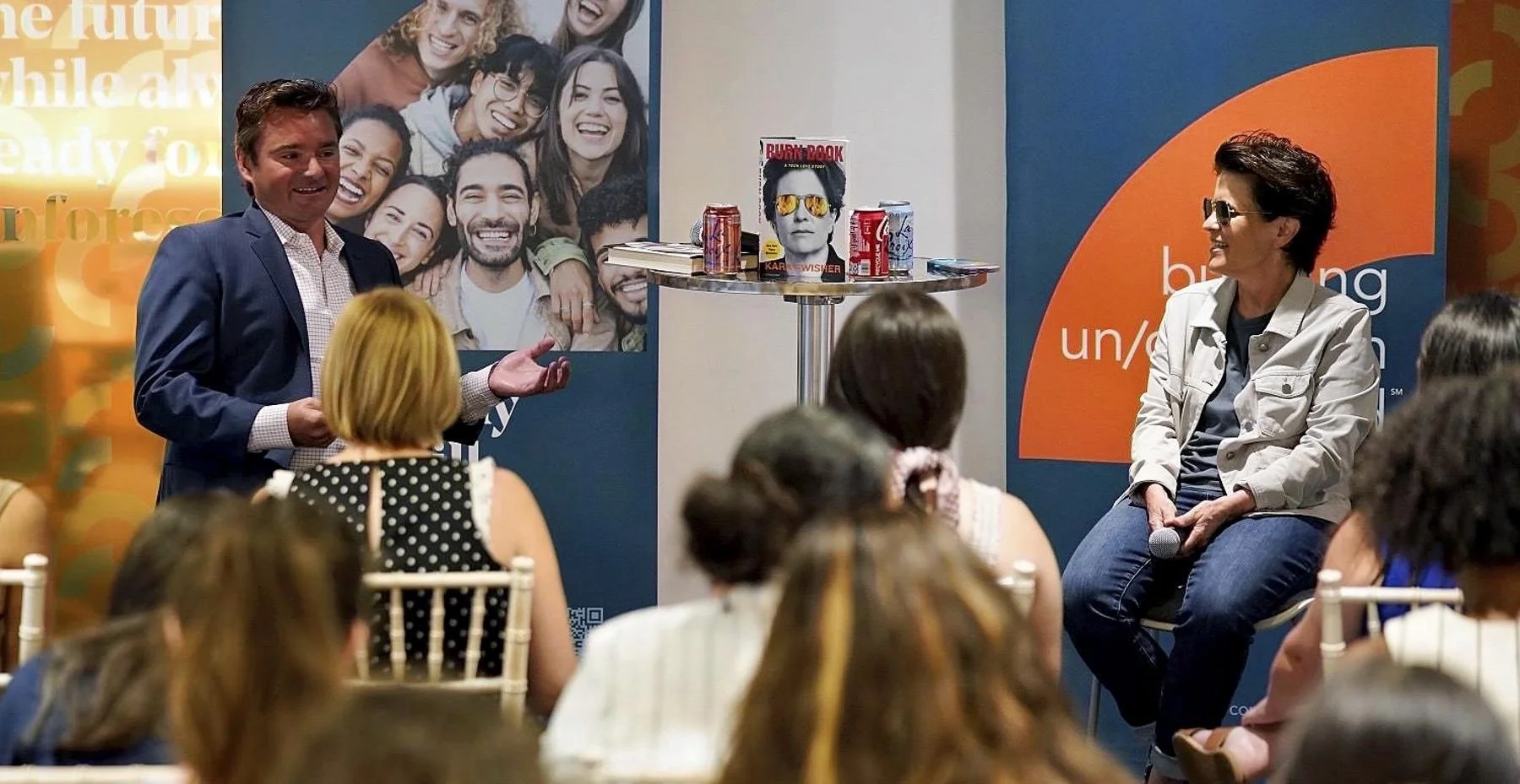 A man in a suit speaks to an audience while a woman in casual attire sits on a stool holding a microphone. Behind them are promotional banners and a table with cans and a book.