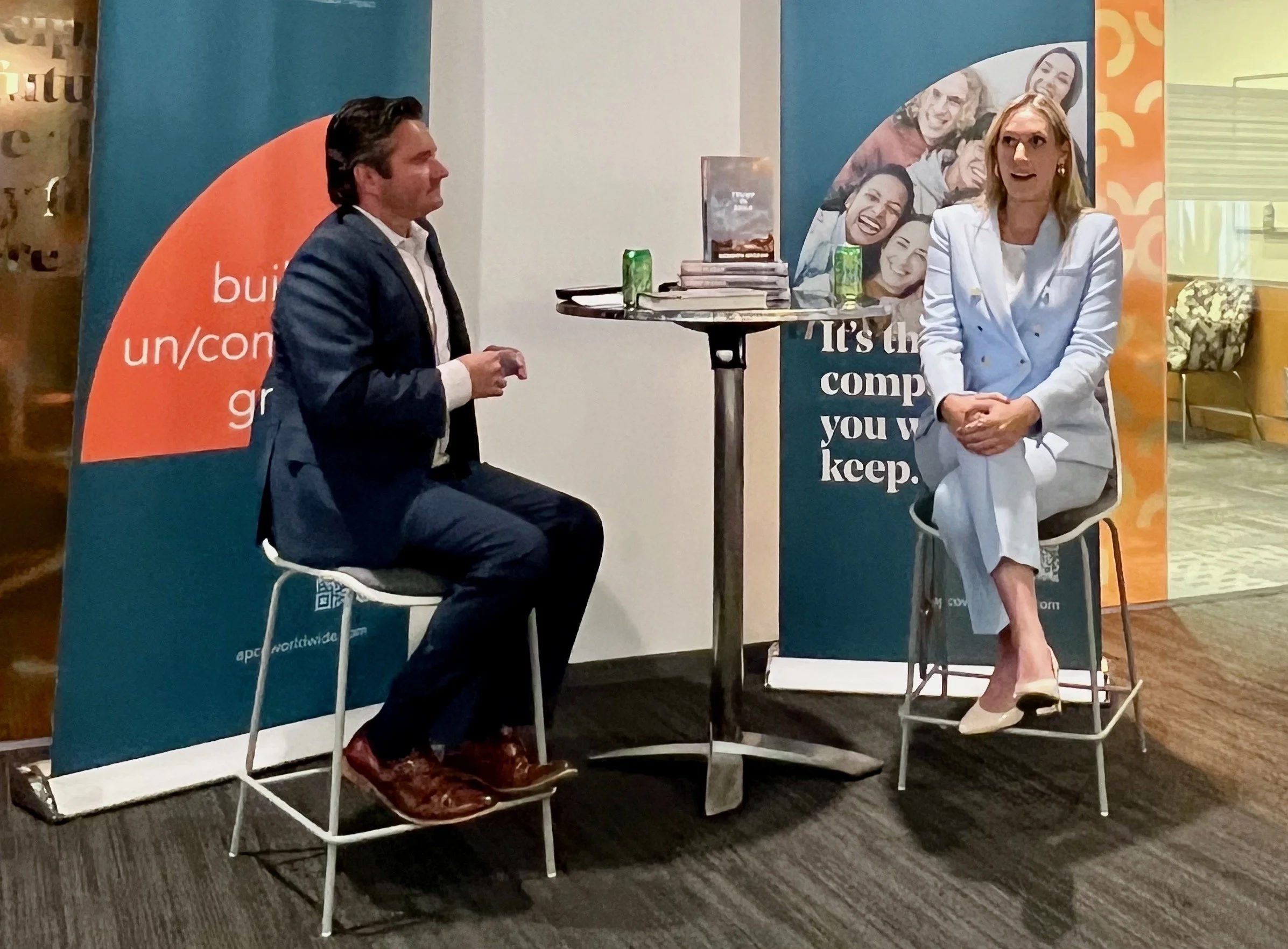 Two people sitting on high stools in a discussion setting, with books and drinks on a small table between them. They are in an office environment with branded banners in the background.