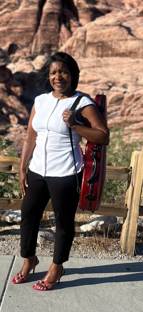 Valencia Thevenin standing outdoors near rocks, carrying violin on her back, wearing a white blouse, and black pants..
