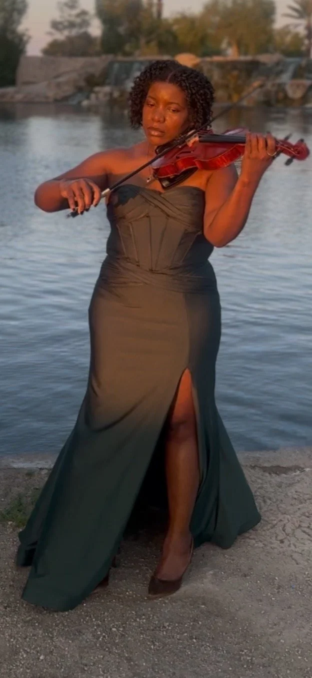 Valencia Thevenin in a dark green dress, standing by a body of water, playing a violin.