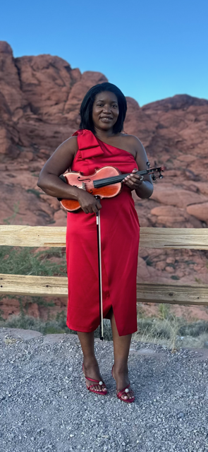 Valencia Thevenin in a red dress holding a violin and bow, standing outdoors near a wooden fence with rock formations and blue sky in the background.