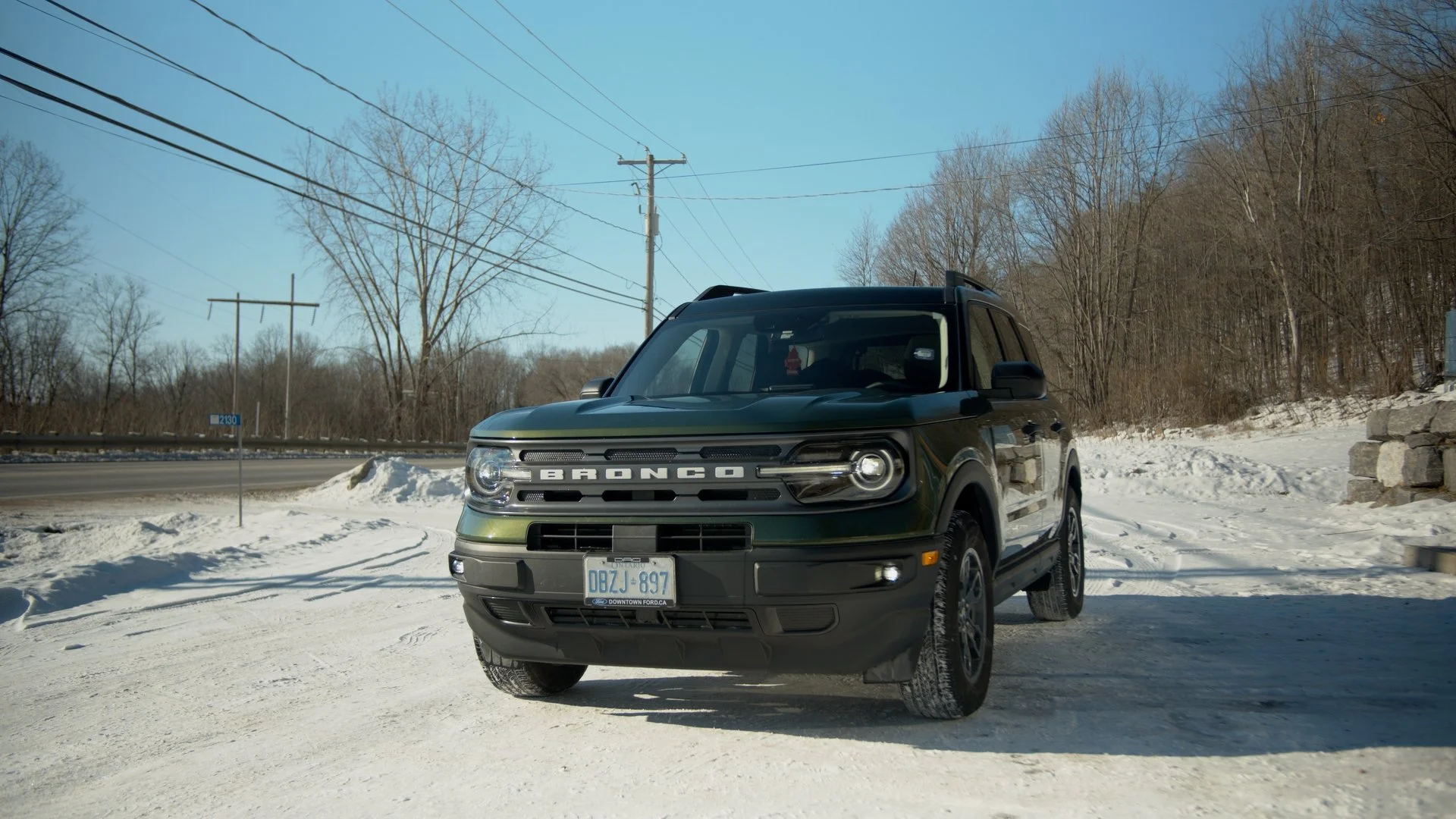 Green Ford Bronco parked on snow-covered road with trees in background.