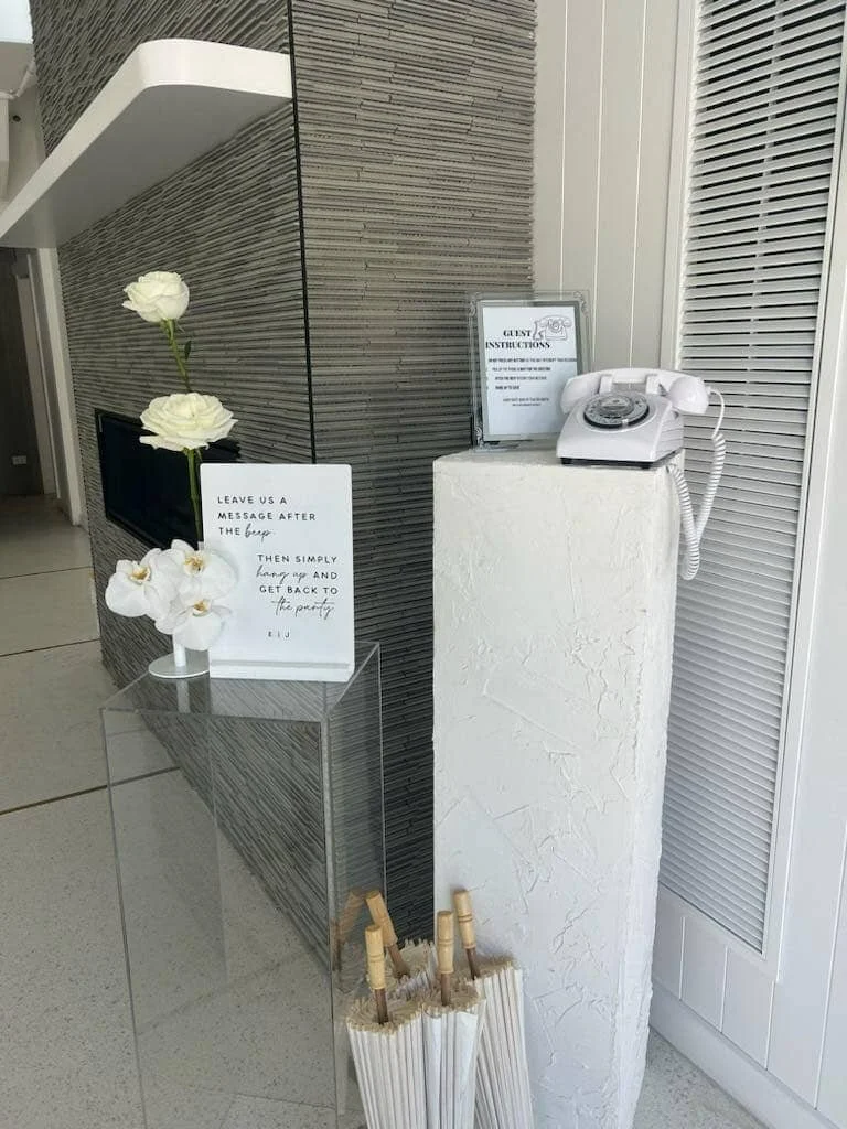 A premium white vintage rotary audio guestbook phone styled on a textured white plinth with modern acrylic signage and fresh white orchids at an upscale event venue