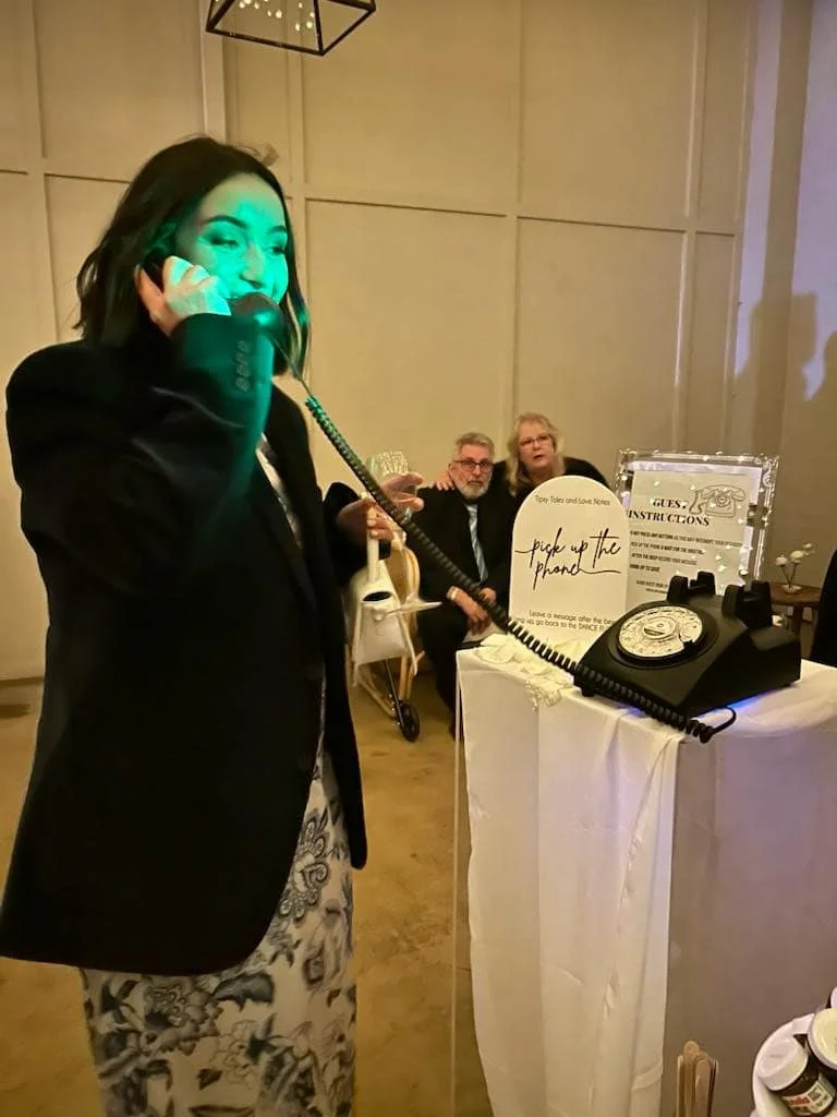 A smiling female event guest holds the receiver and speaks into a black vintage rotary audio guestbook phone styled on a white draped table next to a custom instruction sign