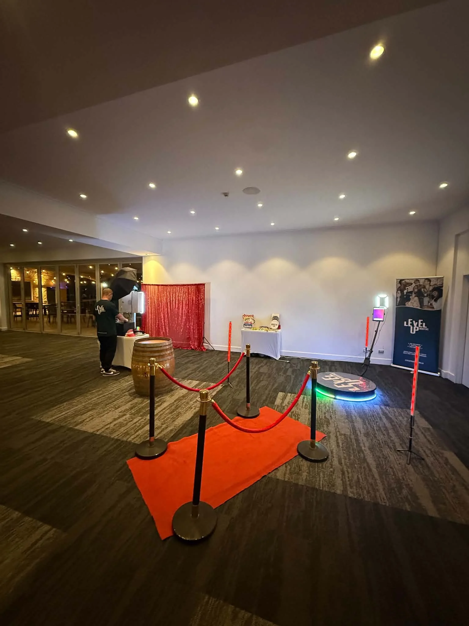 A wide shot of a premium Level Up Booths event setup featuring a VIP red carpet entry with gold stanchions, an open-air photo booth with a red curtain, and a glowing 360 video platform in a large venue
