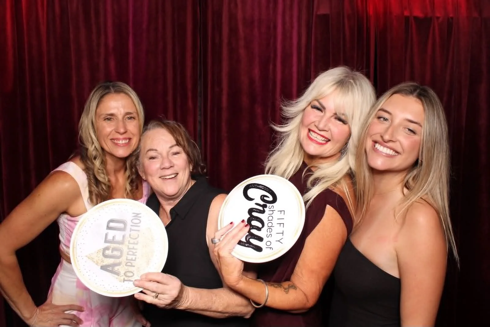 Four smiling women pose happily with fun props, including "AGED TO PERFECTION" and "FIFTY SHADES OF CRAZY," in front of a rich, red velvet curtain backdrop, demonstrating Level Up Booths' open-air photo booth quality