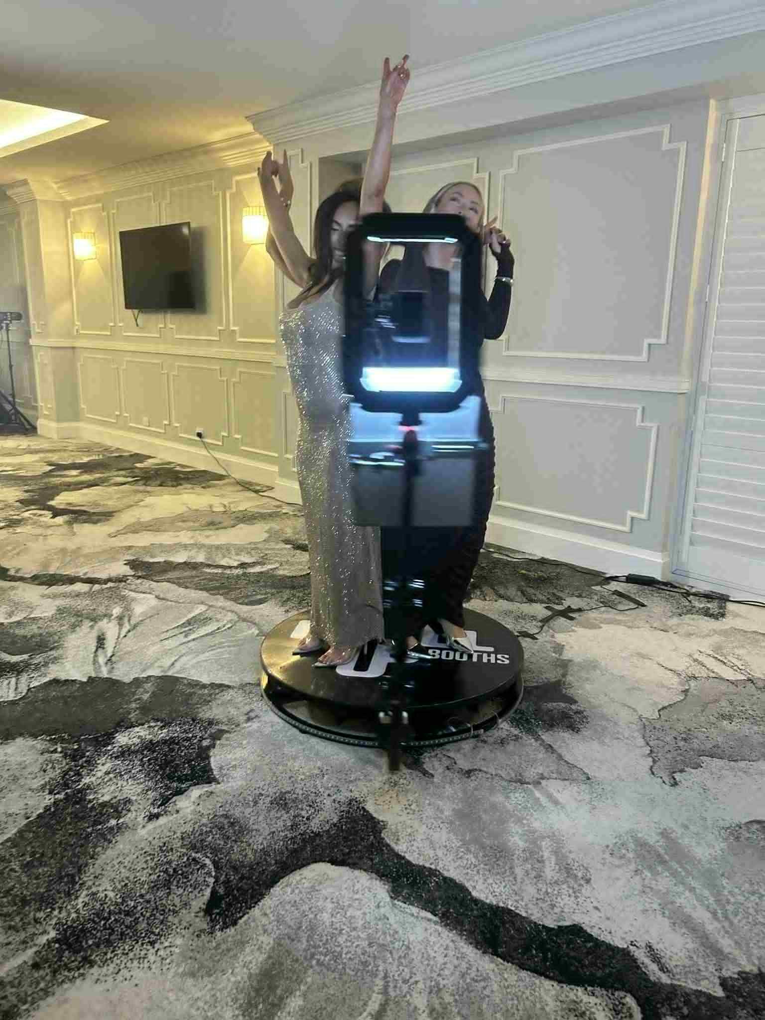 Two glamorous event guests posing with their arms raised on a Level Up Booths branded 360 video platform as the spinning camera arm captures high-energy video footage inside a premium function room