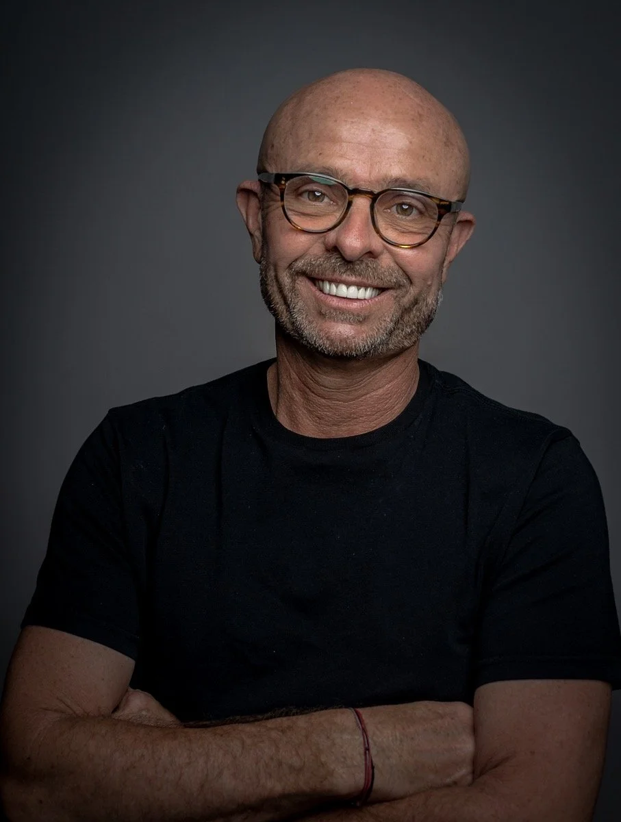 A portrait of middle-aged man with light skin, bald head, wearing glasses, smiling, with his arms crossed, dressed in a black t-shirt, against a dark gray background.