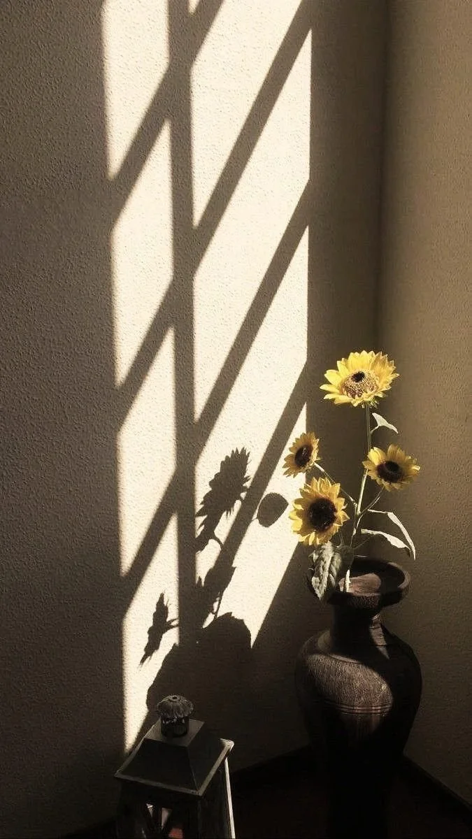 Sunflowers in a vase casting shadows on a textured wall with sunlight, next to a small table with a decorative item.
