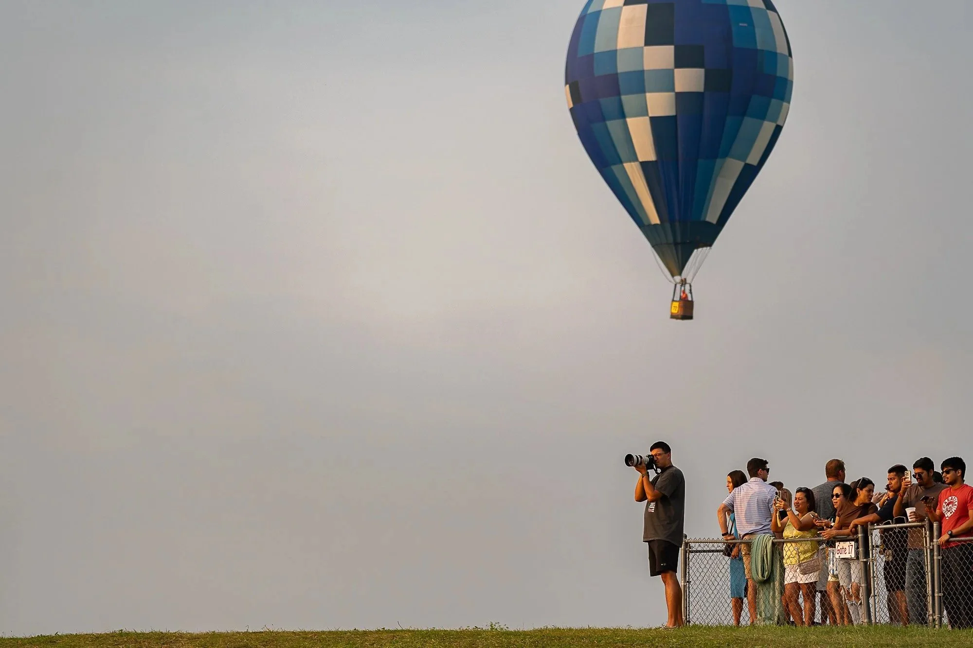 A hot air balloon in a patchwork of blue, beige, and dark colors flying in the sky. Below, a crowd of people stands behind a fence, some taking photos, with a person in the foreground holding a camera with a large lens.