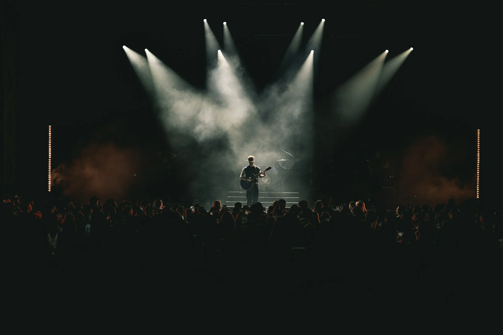 A musician performing on stage with a guitar under bright lights, with an audience watching in the dark.