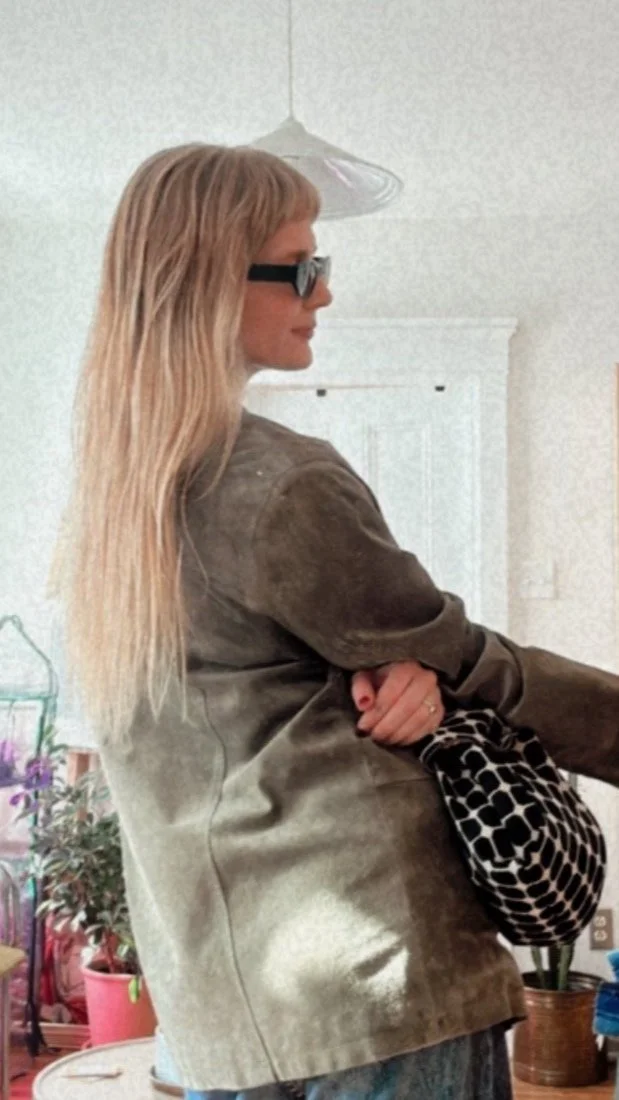 Une personne aux cheveux longs portant des lunettes de soleil et une veste, tenant un sac à motifs noirs et blancs. En arrière-plan, des plantes et un intérieur lumineux.