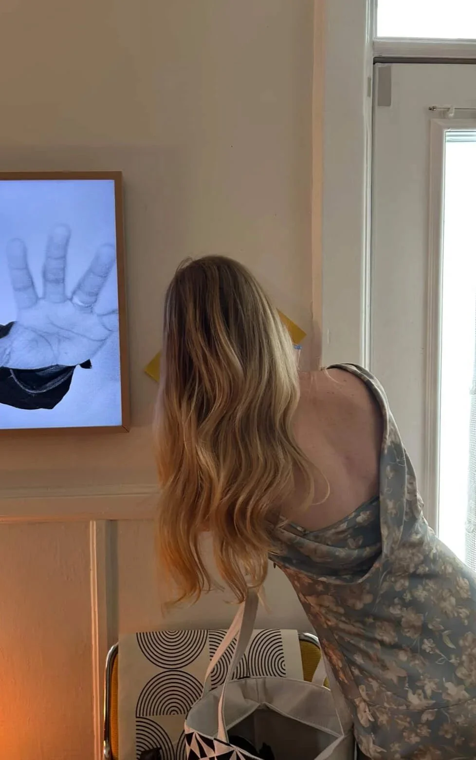 Une femme avec de longs cheveux blonds regarde une télévision affichant une personne portant des lunettes blanches et faisant un geste avec la main.