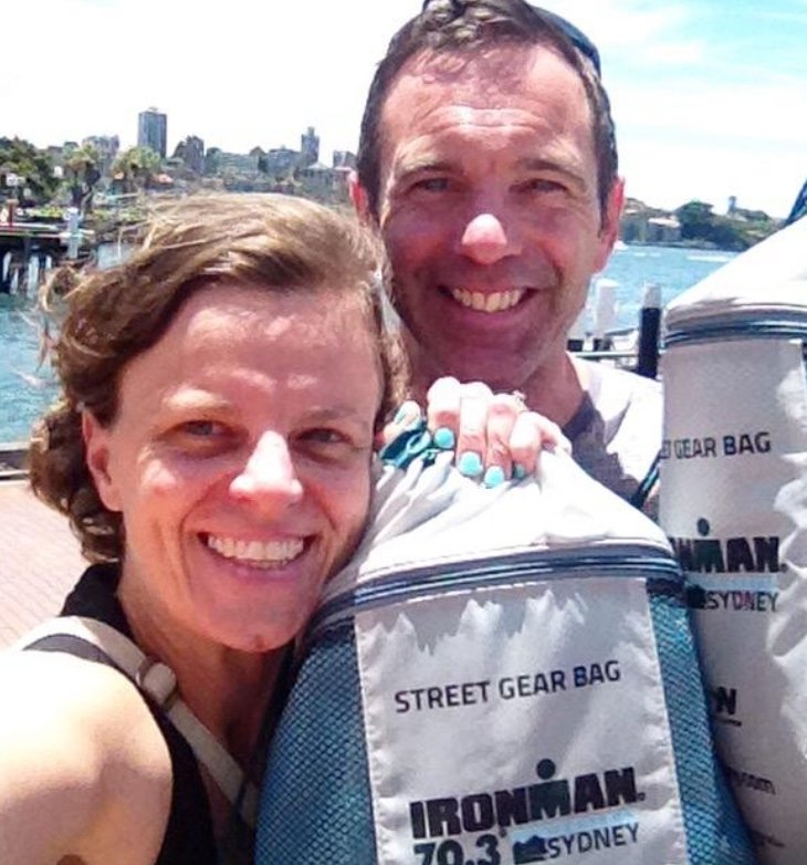 Two people smiling with Ironman 70.3 Sydney gear bags in a sunny outdoor setting.