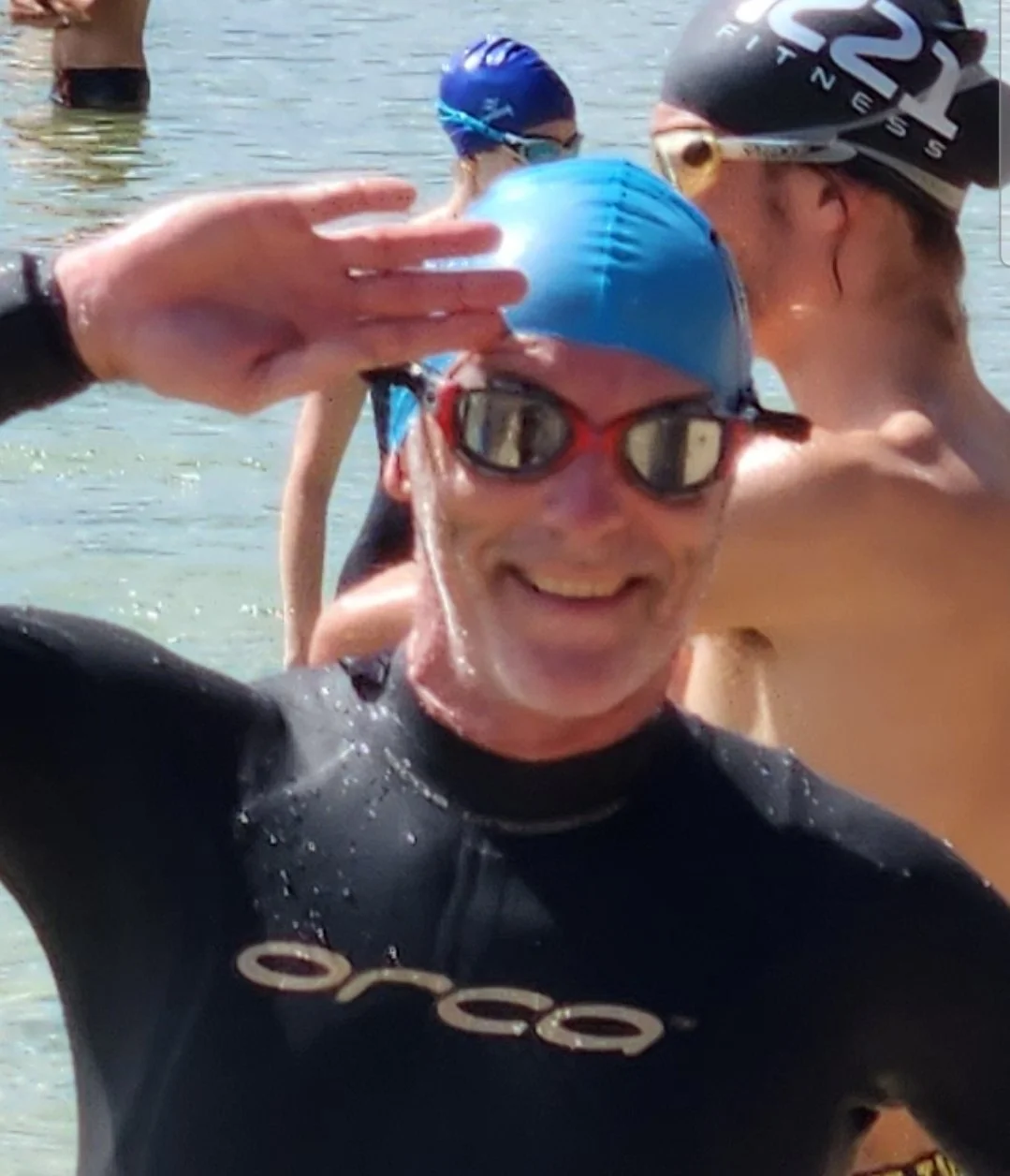 Group of swimmers in wetsuits and swim caps at a beach, one saluting the camera.
