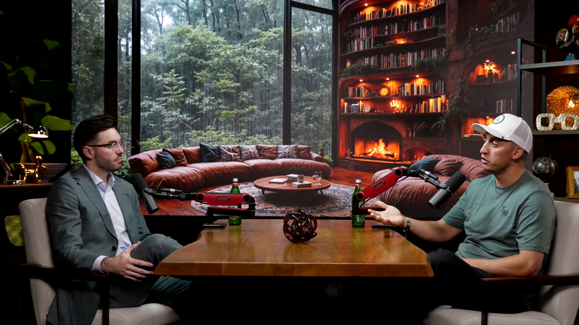 Two men seated at a table in a recording studio with a forest scene background, equipped with microphones, bottles, and a decorative object.